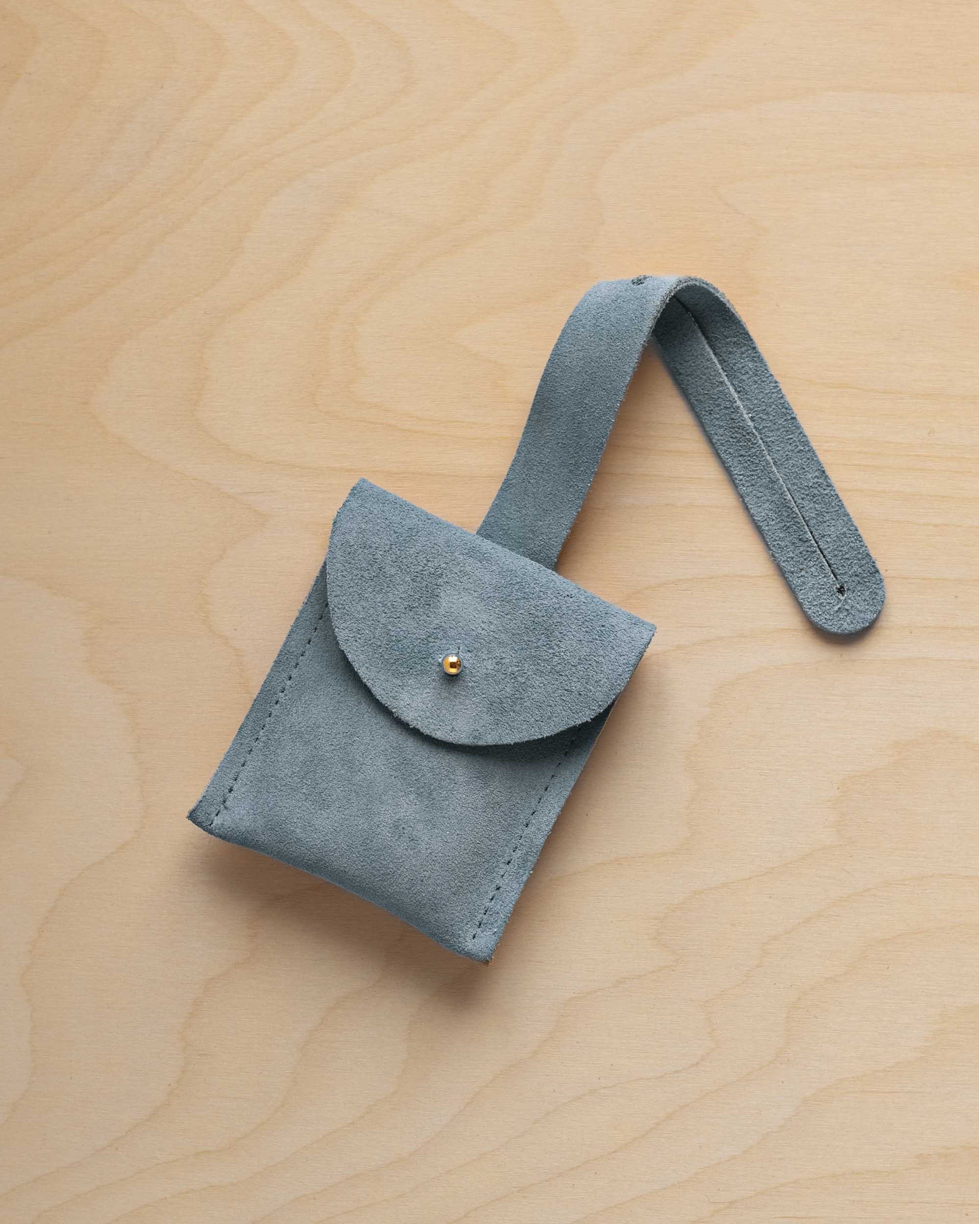 A small dusly blue suede pouch with a flap closure and a matching strap, resting on a light wood grain surface.