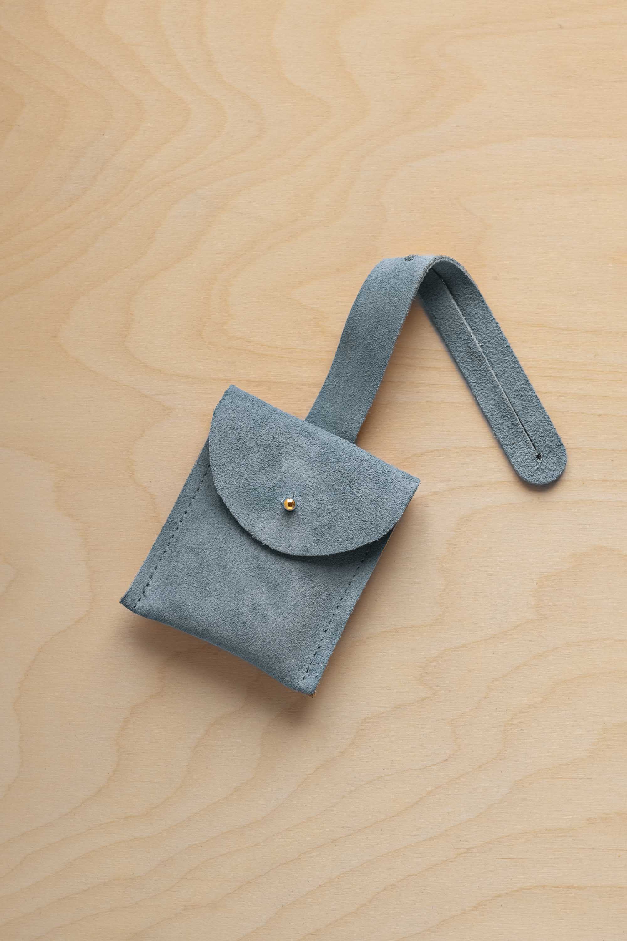 A small dusly blue suede pouch with a flap closure and a matching strap, resting on a light wood grain surface.