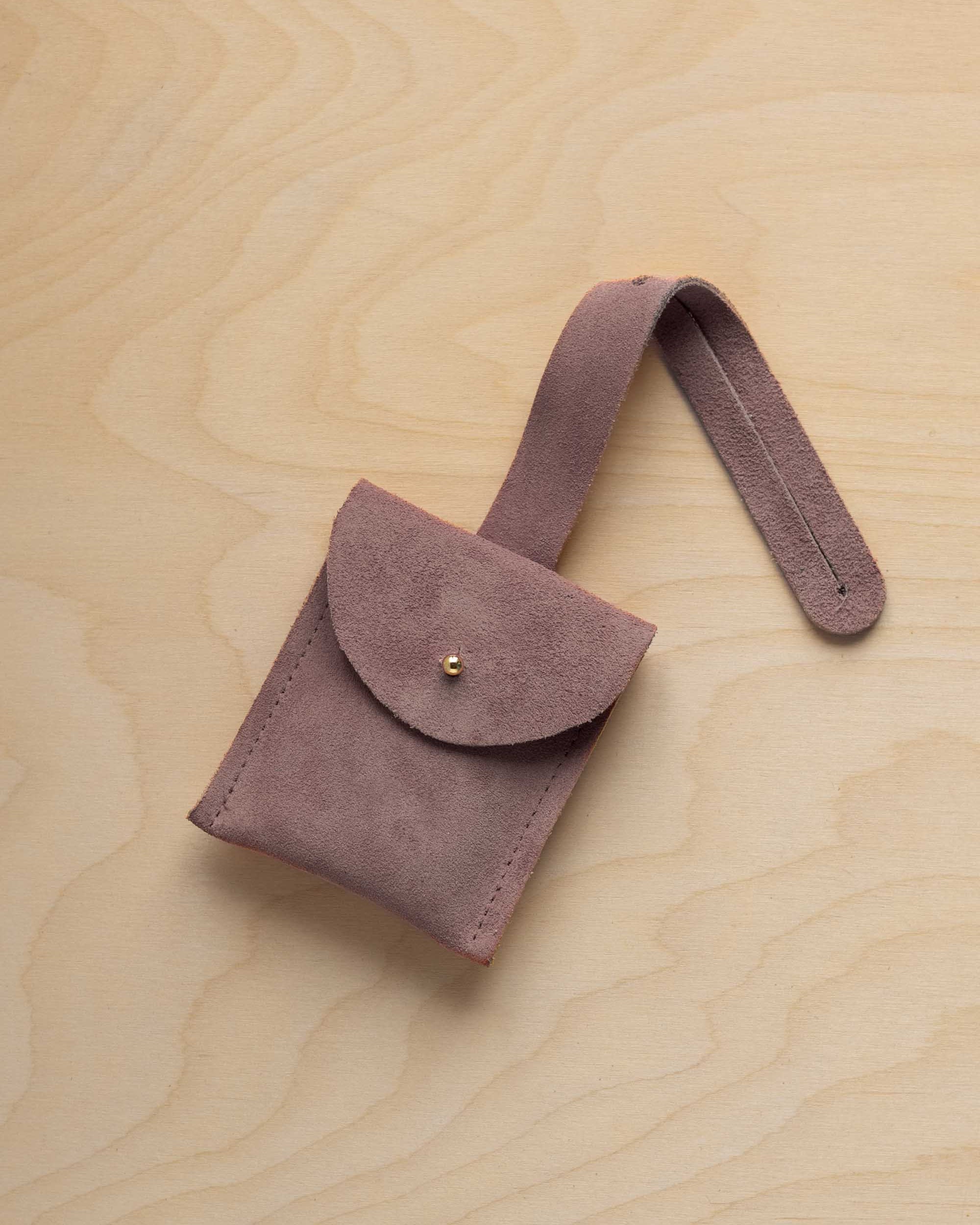 A small dusty mauve grey suede pouch with a flap closure and a matching strap, resting on a light wood grain surface.