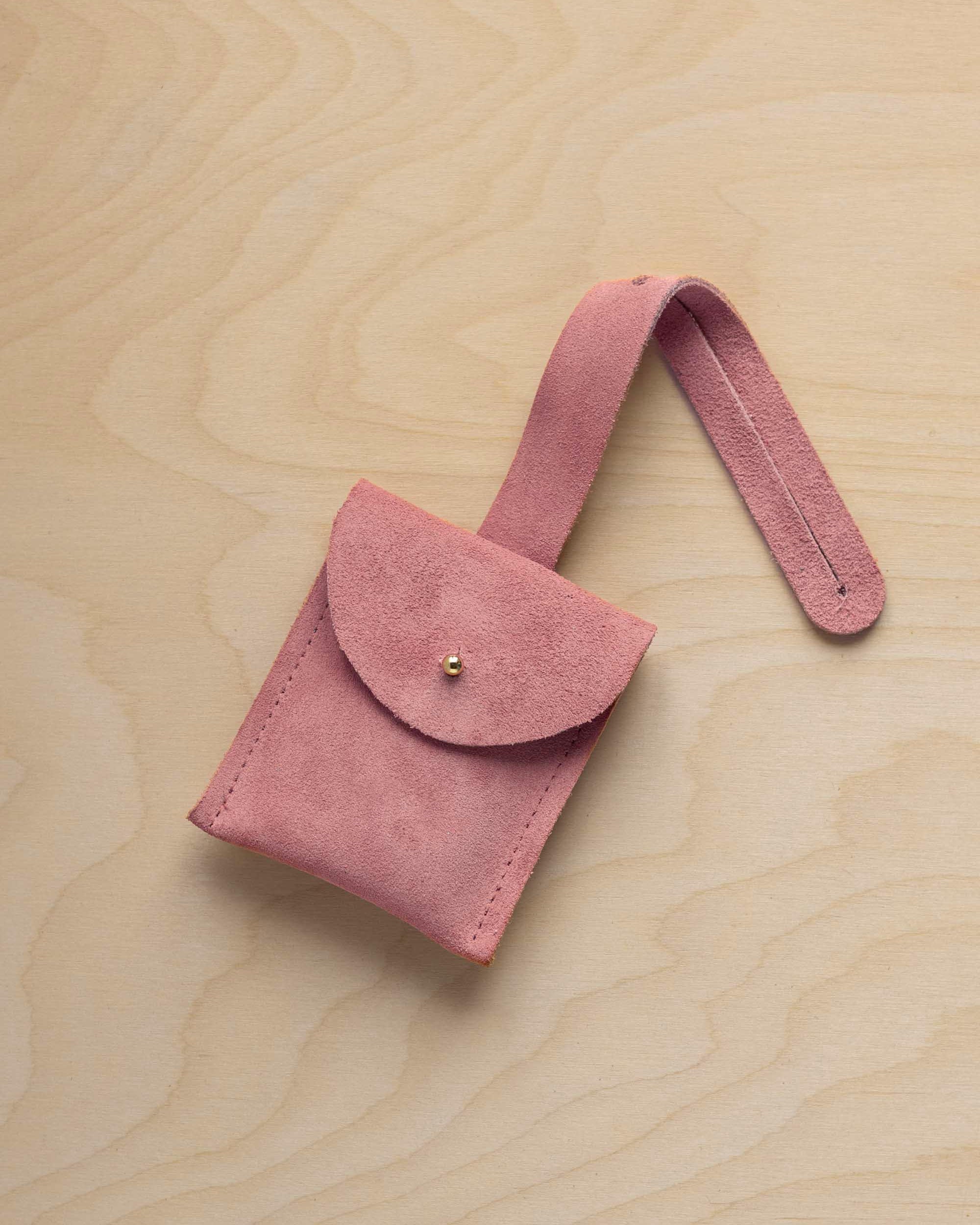 A small dusty rose pale pink coloured suede pouch with a flap closure and a matching strap, resting on a light wood grain surface.