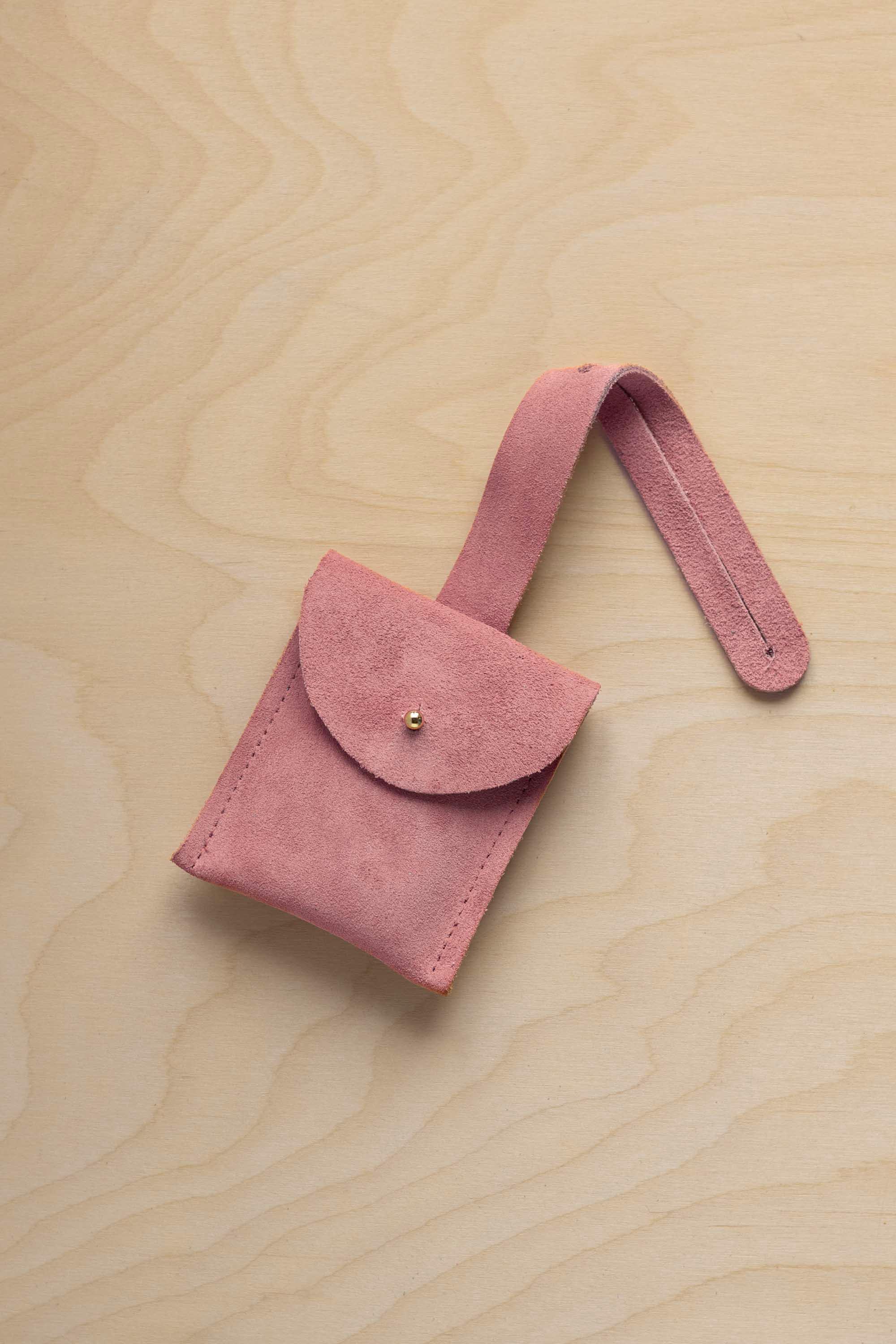 A small dusty rose pale pink coloured suede pouch with a flap closure and a matching strap, resting on a light wood grain surface.