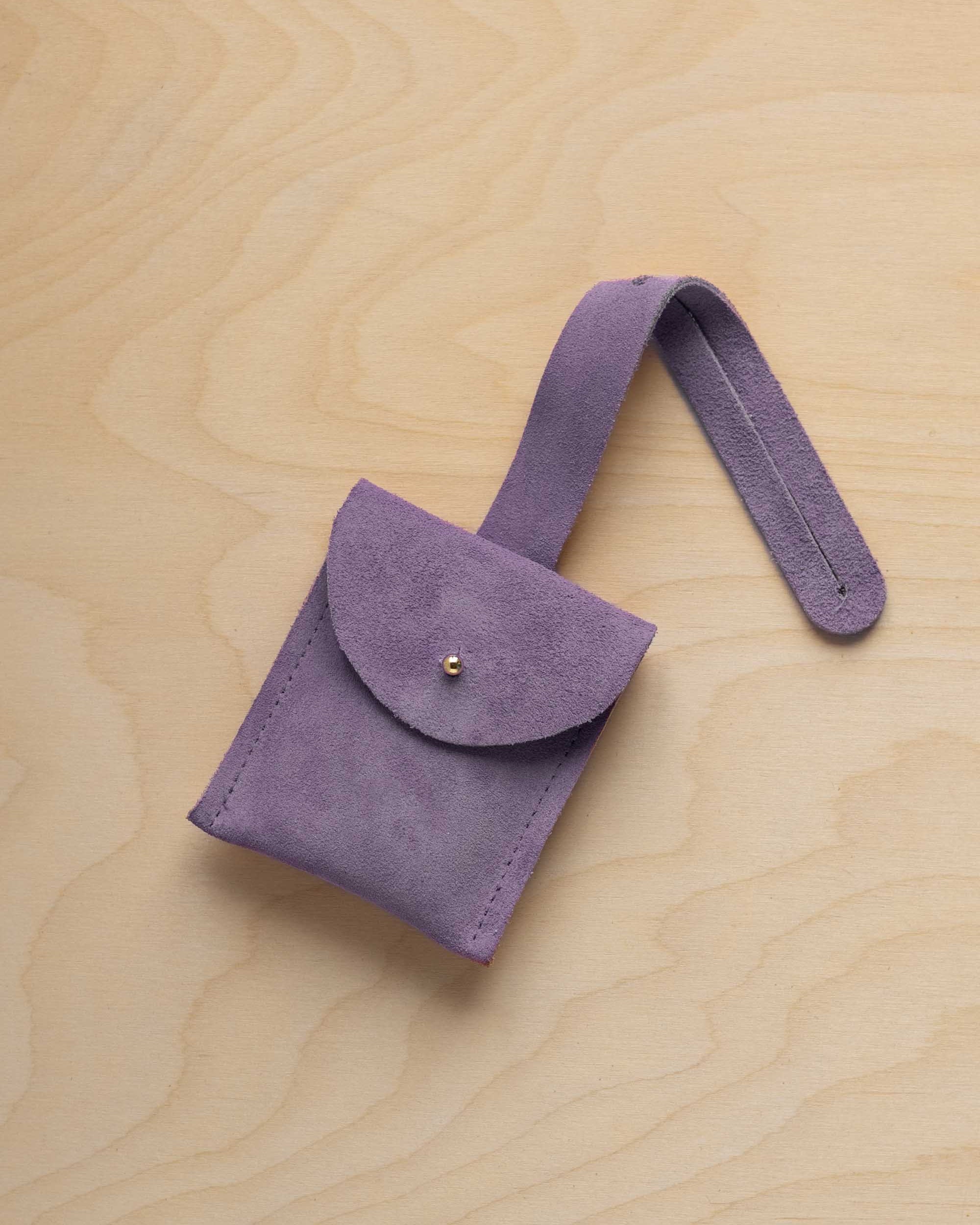 A small lavender coloured suede pouch with a flap closure and a matching strap, resting on a light wood grain surface.