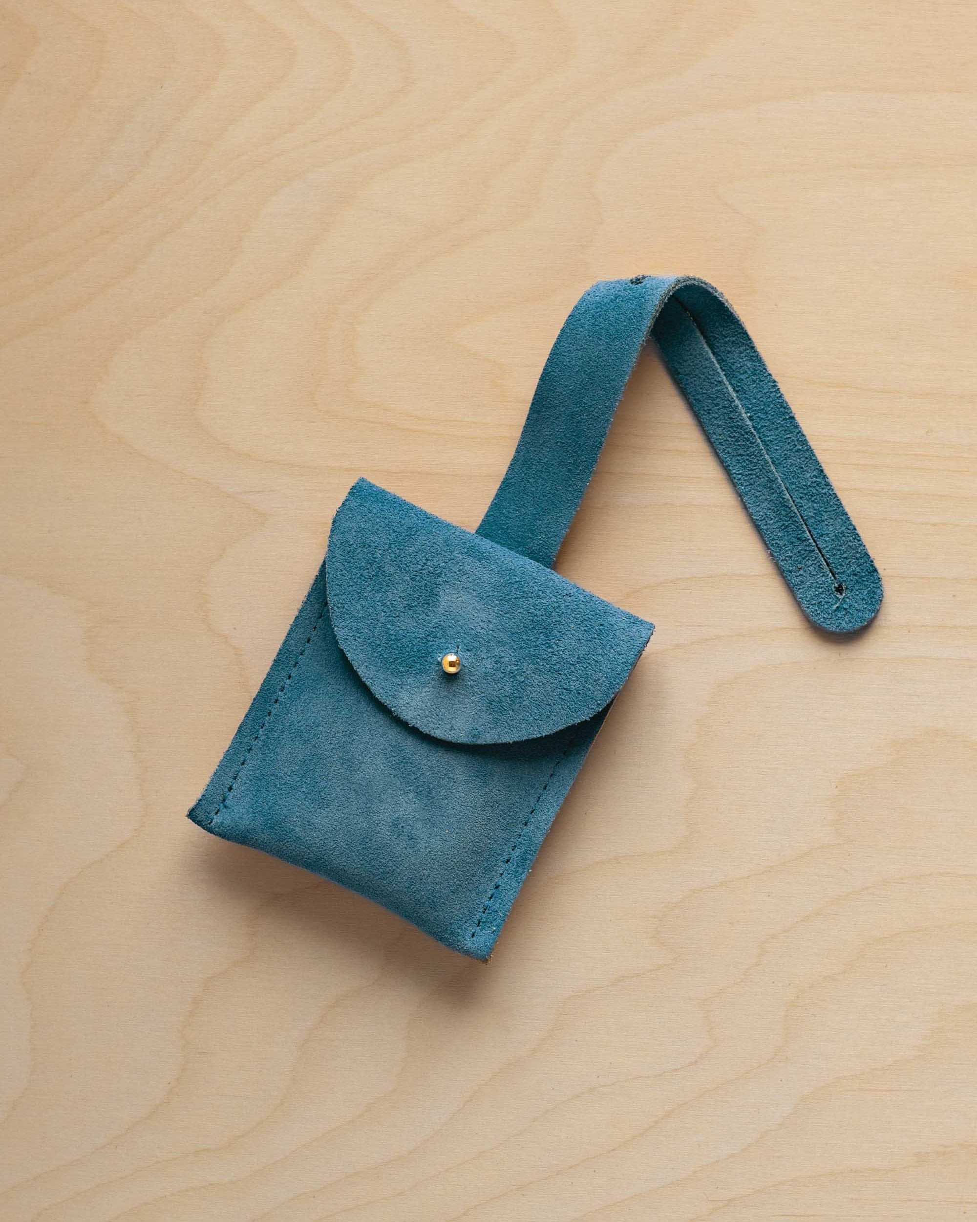 A small marine teal suede pouch with a flap closure and a matching strap, resting on a light wood grain surface.