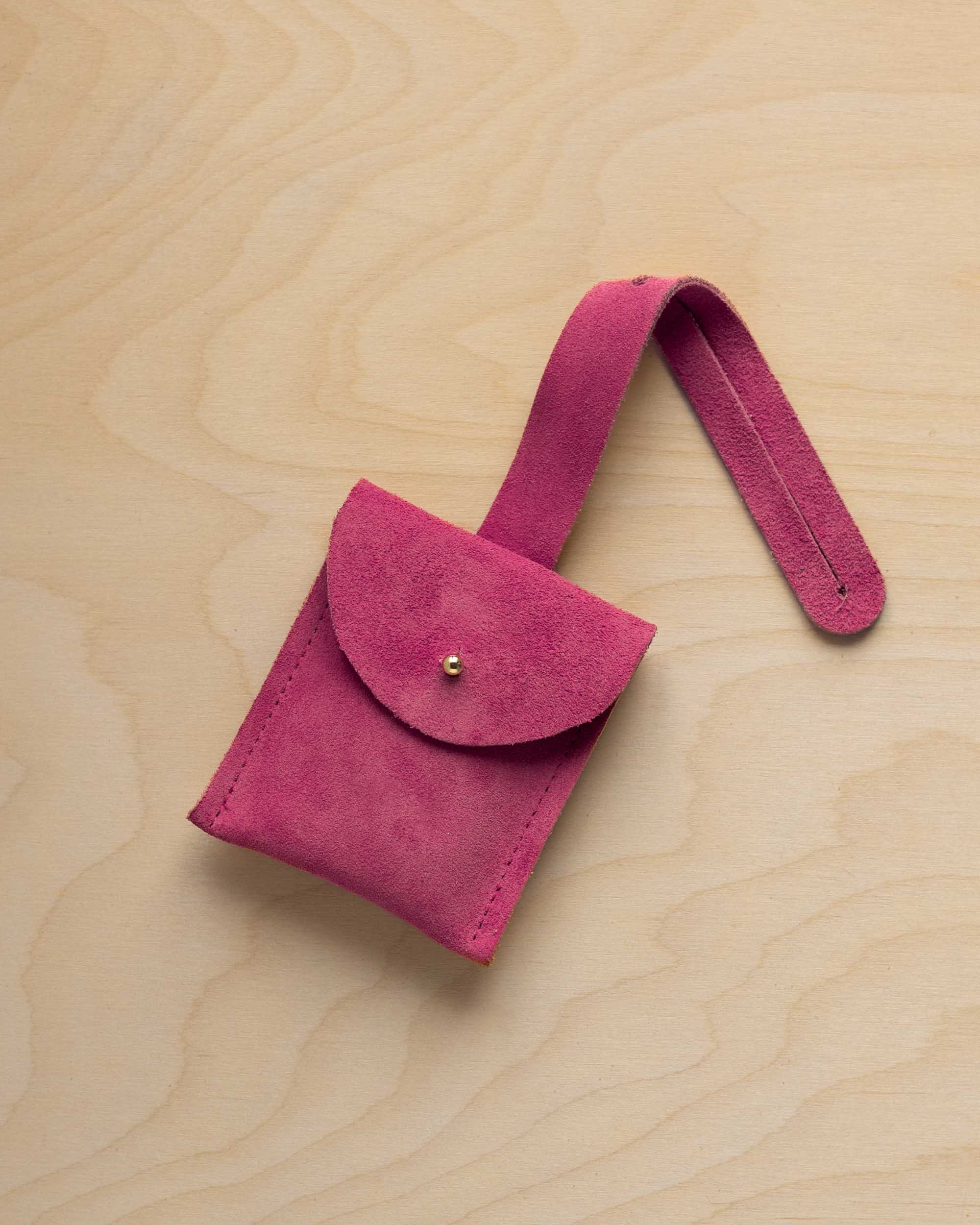 A small raspberry pink suede pouch with a flap closure and a matching strap, resting on a light wood grain surface.