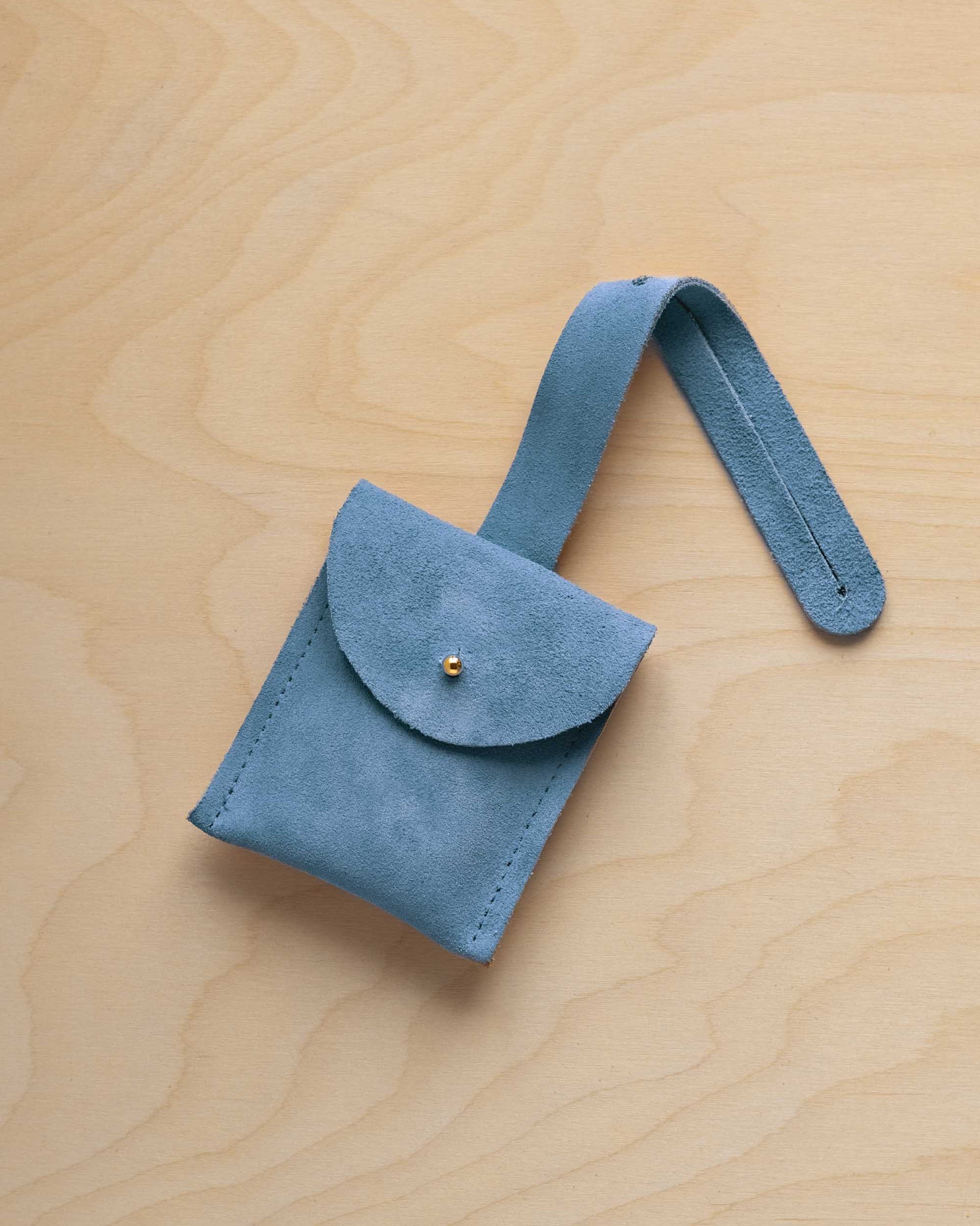 A small sky blue suede pouch with a flap closure and a matching strap, resting on a light wood grain surface.