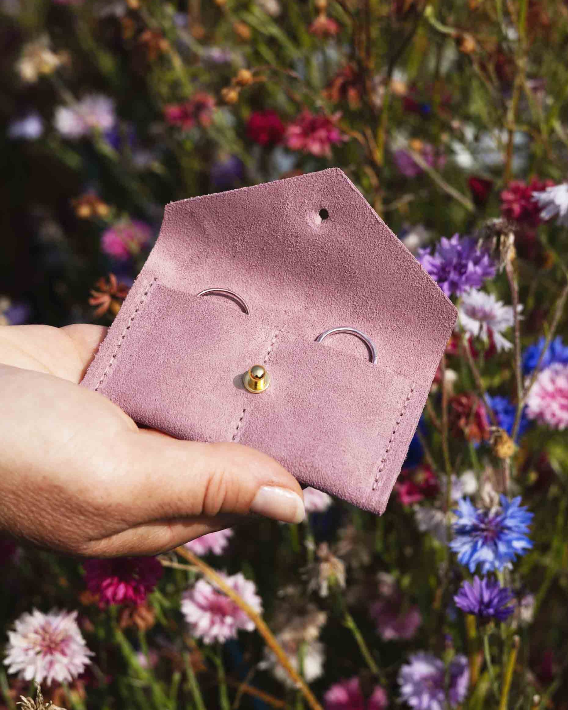 A hand holding a small dusty pink leather pouch with a gold button, set against a blurred background of colourful wildflowers.