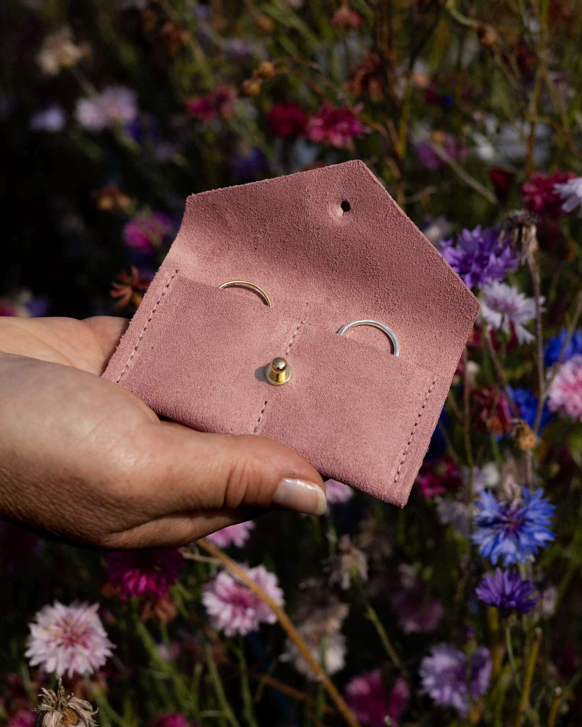 A hand holding a small dusty pink leather pouch with a gold button, set against a blurred background of colourful wildflowers.