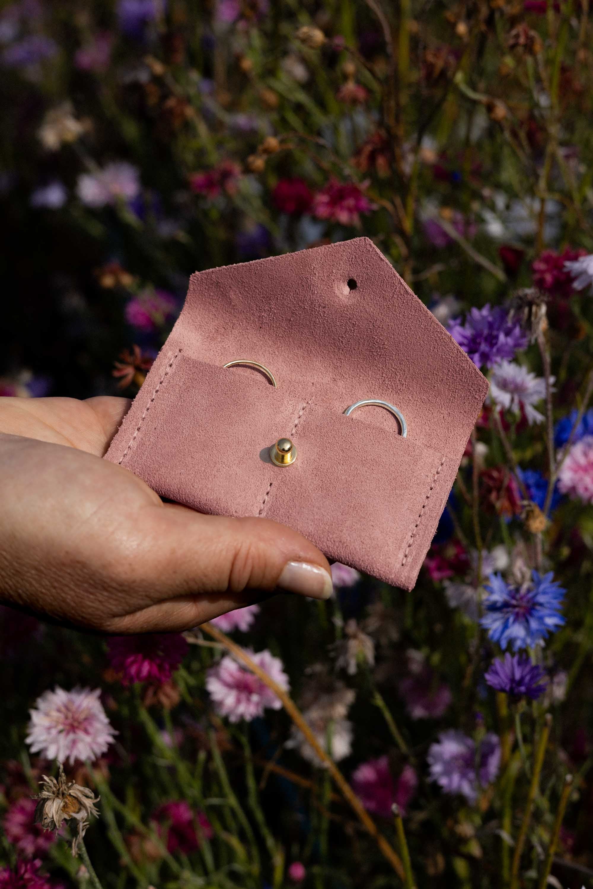 A hand holding a small dusty pink leather pouch with a gold button, set against a blurred background of colourful wildflowers.