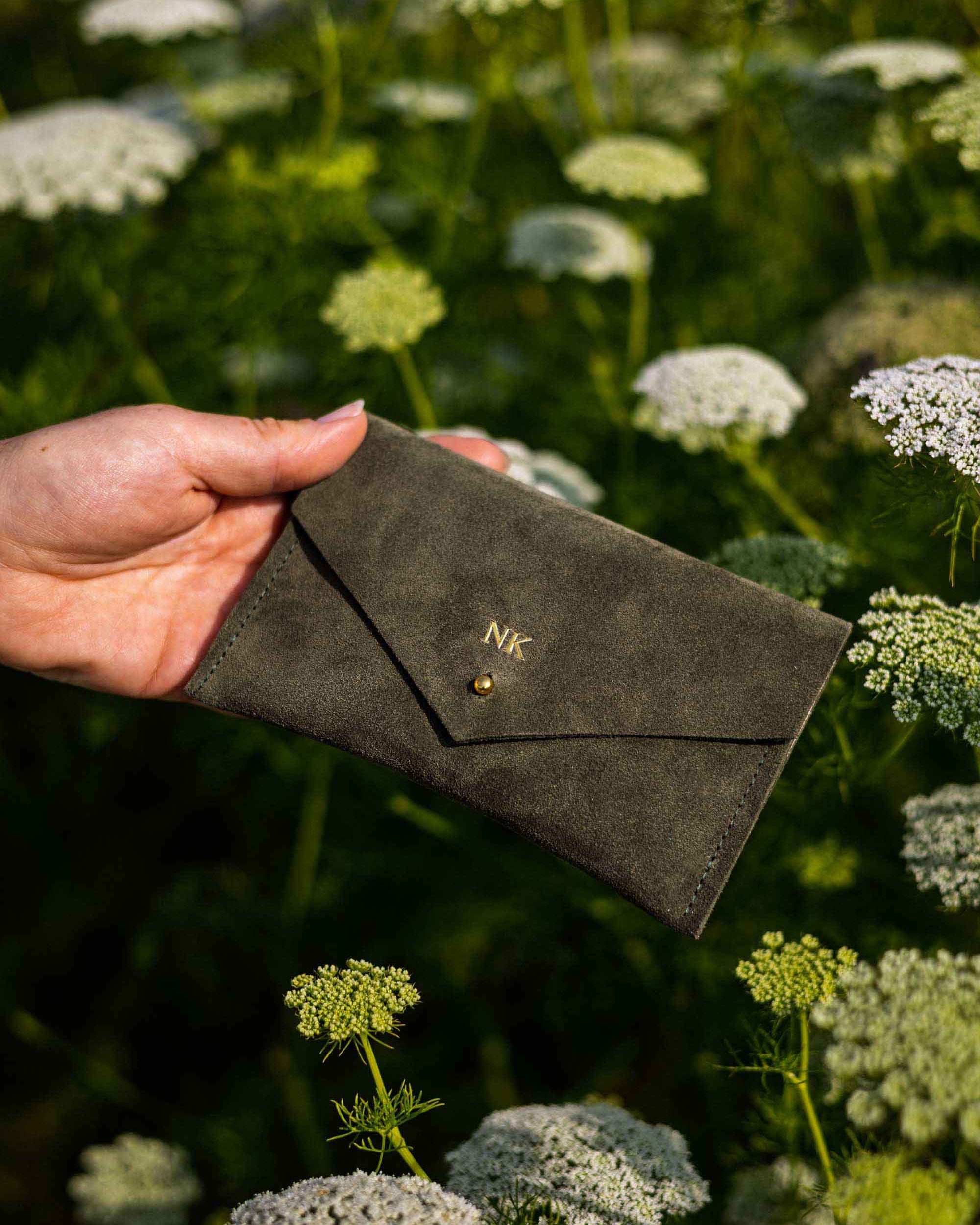 A person's hands holding an open moss or olive green envelope-style wallet with a gold-tone button, set against a blurred background of green foliage and white wildflowers.