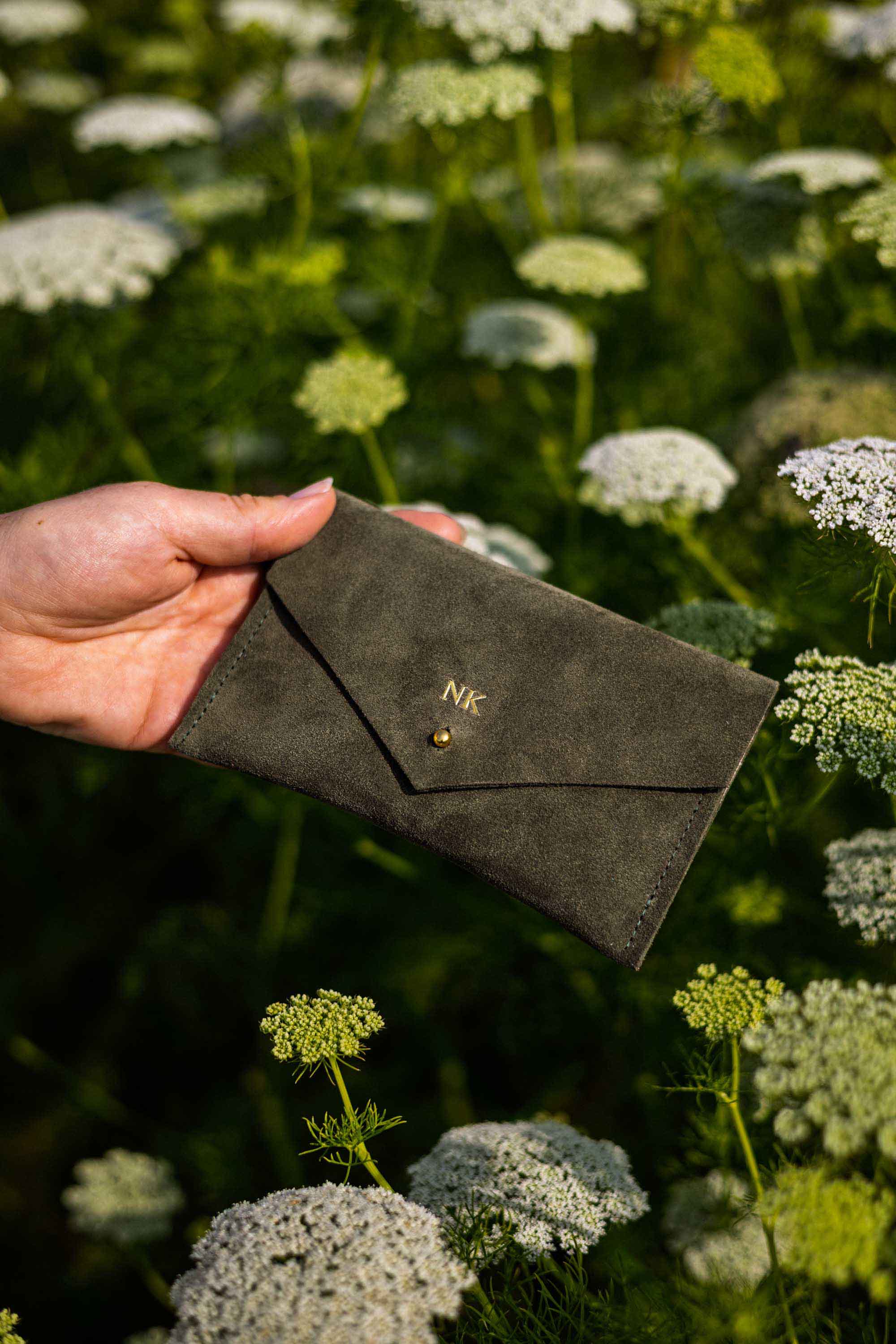 A person's hands holding an open moss or olive green envelope-style wallet with a gold-tone button, set against a blurred background of green foliage and white wildflowers.