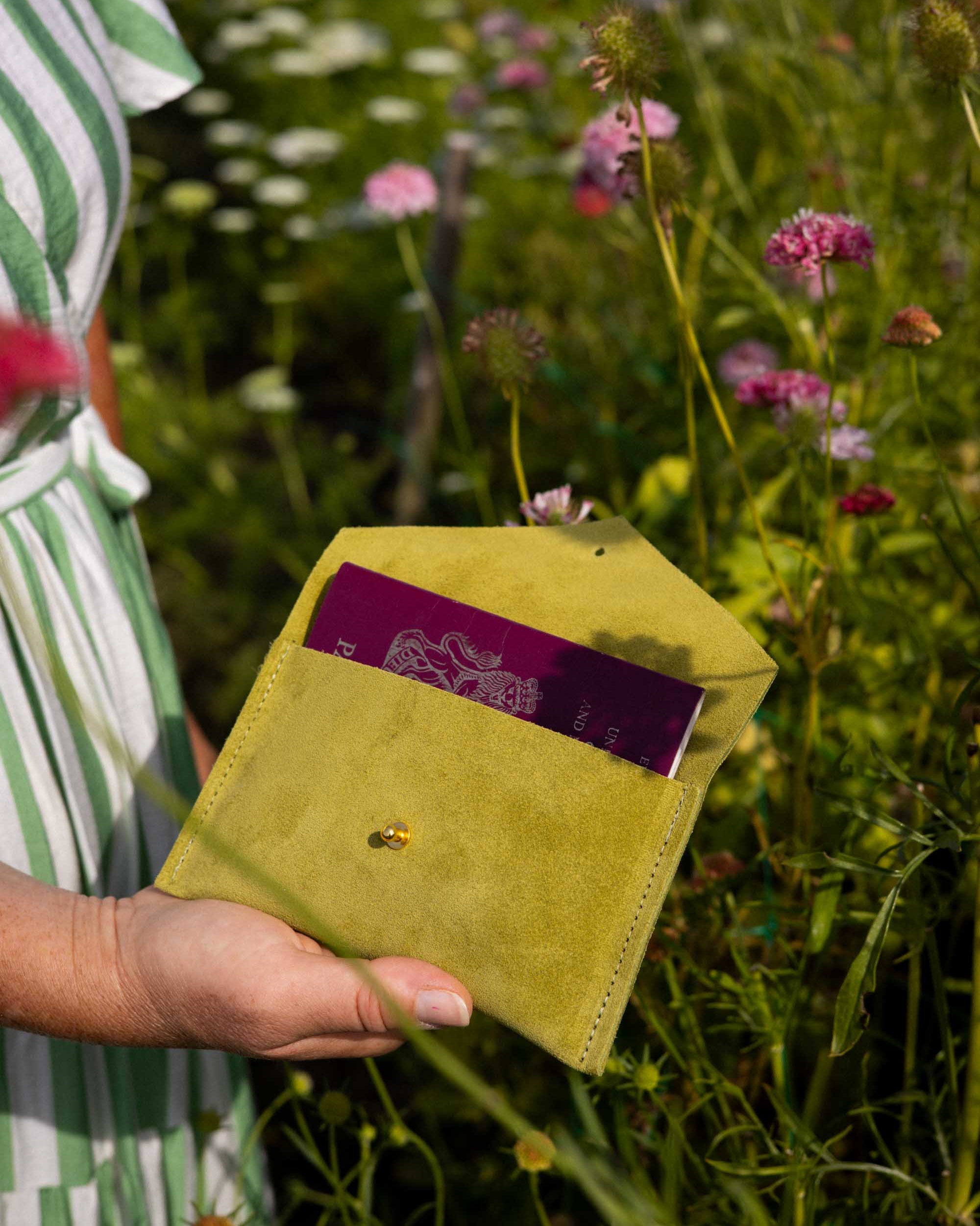 A person outdoors in a flower field holds a small olive-yellow envelope-style pouch with a passport partially inserted. The background is a blurred green and pink wildflower meadow.