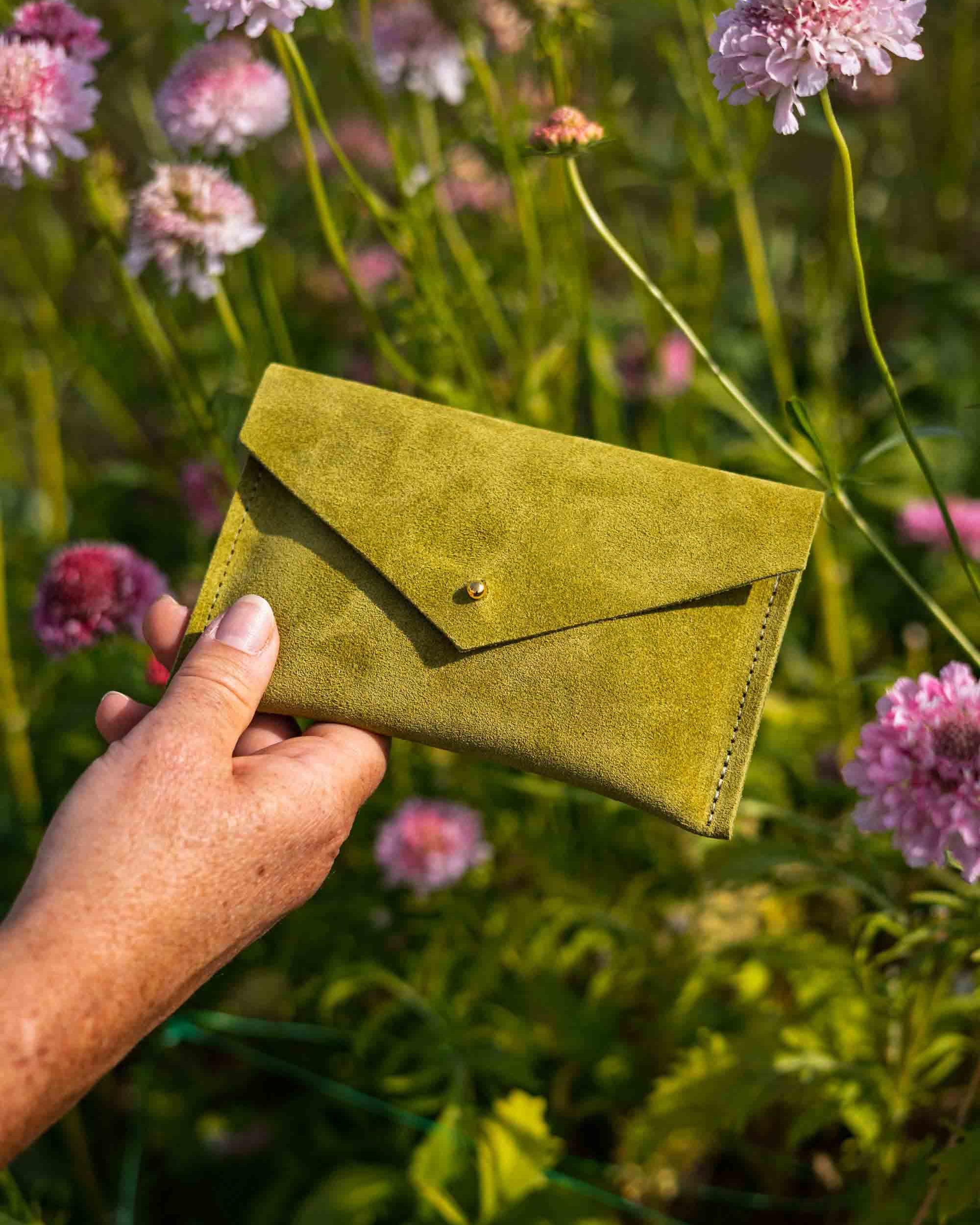 A hand holds a small olive green suede envelope-style wallet or clutch with a button closure, set against a blurred outdoor background of pink flowers and green foliage.