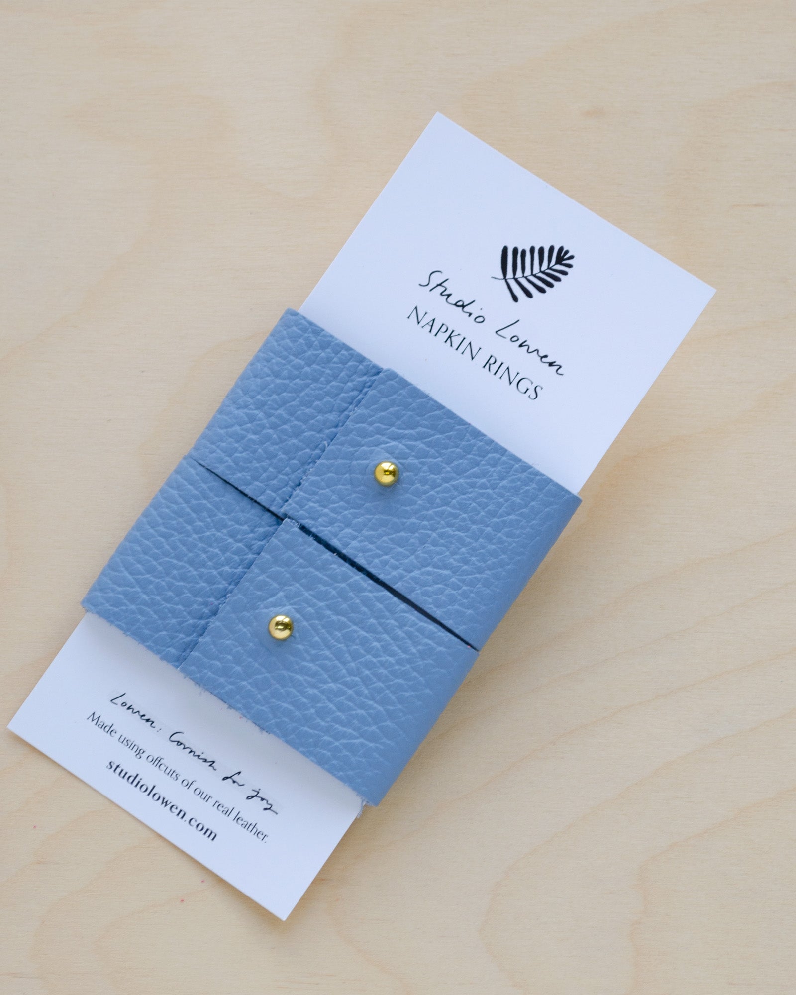 Pair of soft Periwinkle blue leather napkin rings with a gold stud on a white card packaging.