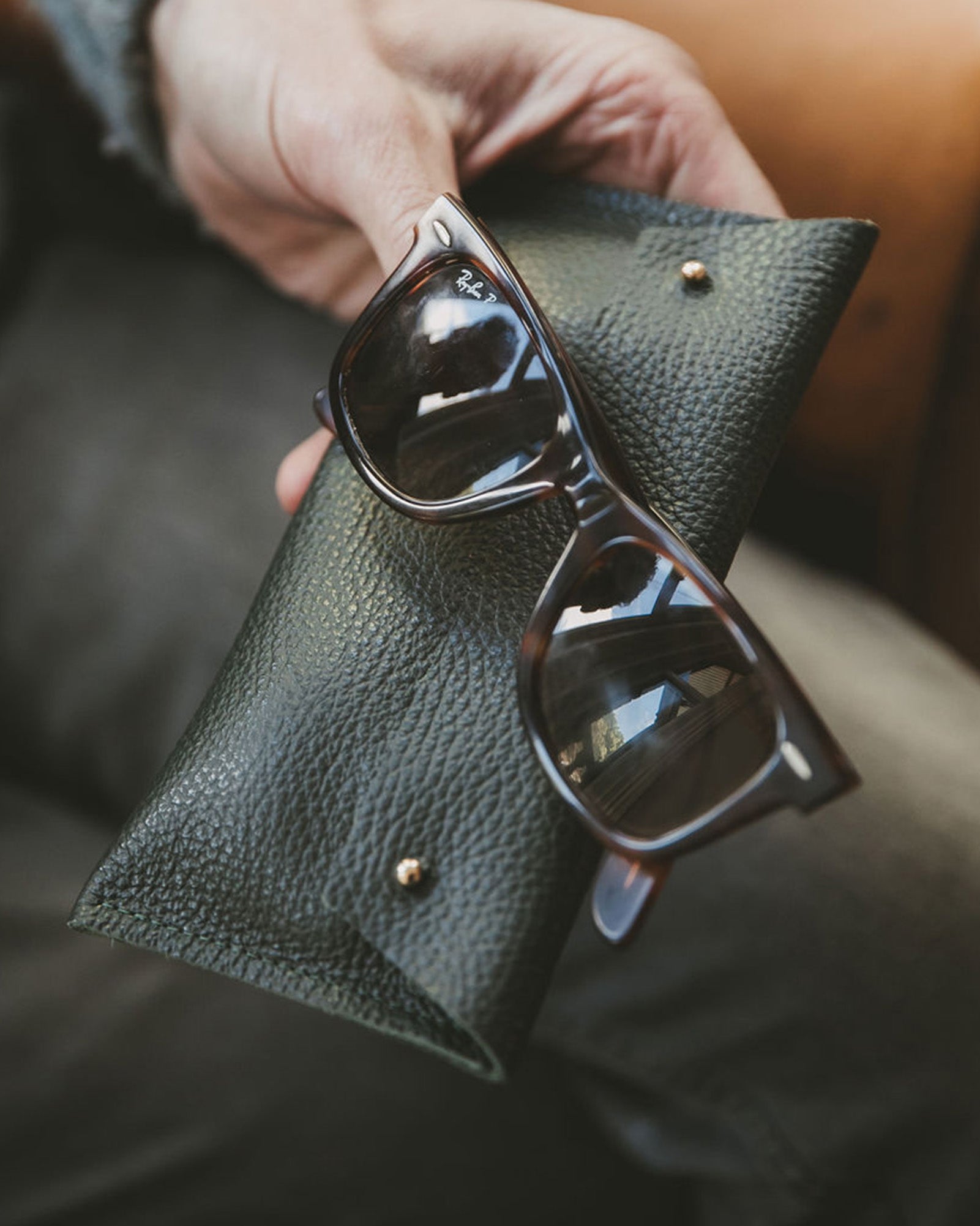 Dark Green leather glasses case in a man's hands with some brown Rayban sunglasses.