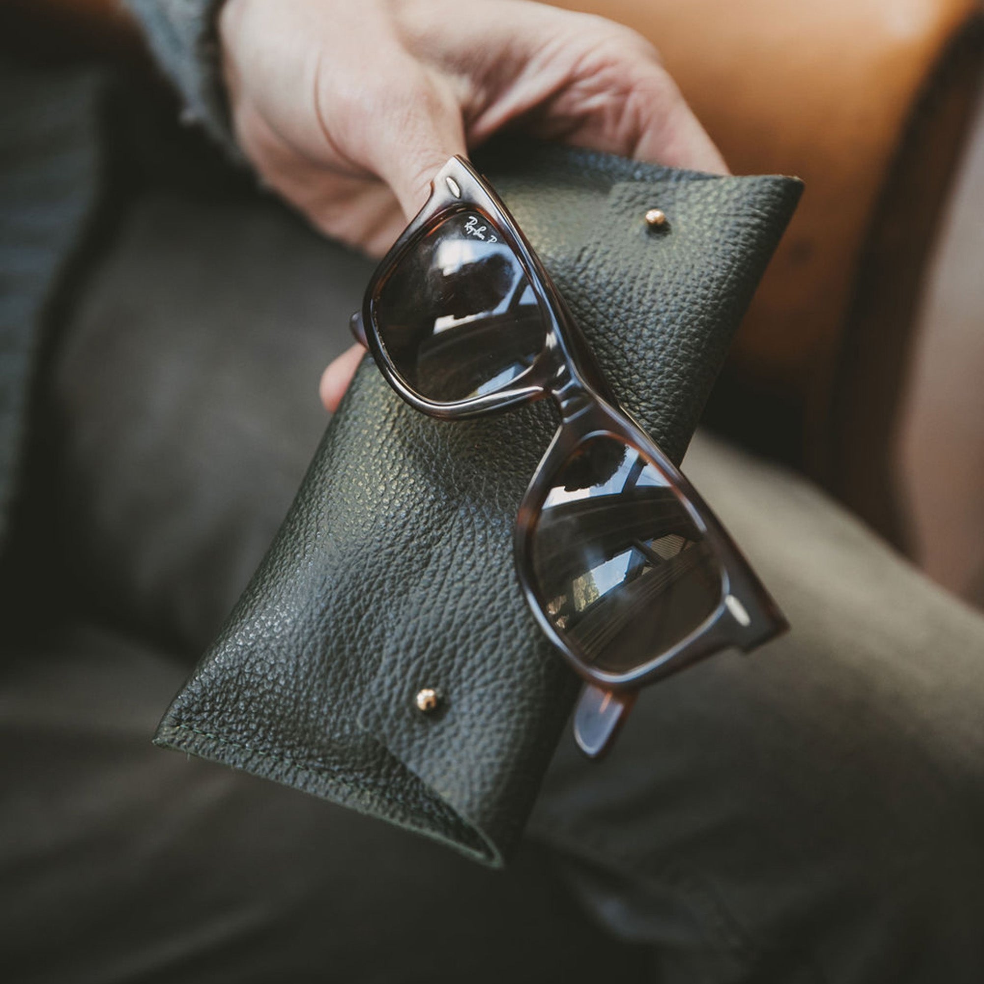 Dark Green leather glasses case in a man's hands with some brown Rayban sunglasses.