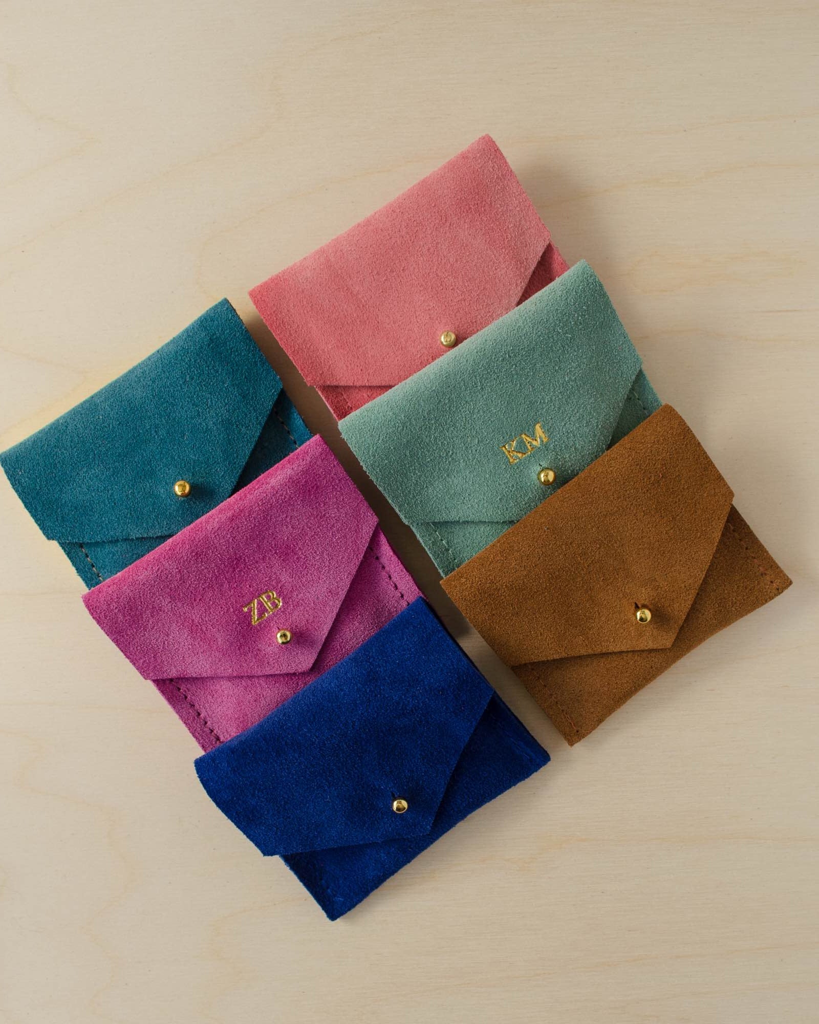 Collection of suede jewellery pouches. Teal, pink, coral, fuchsia, blue and brown options sit on a plywood table.
