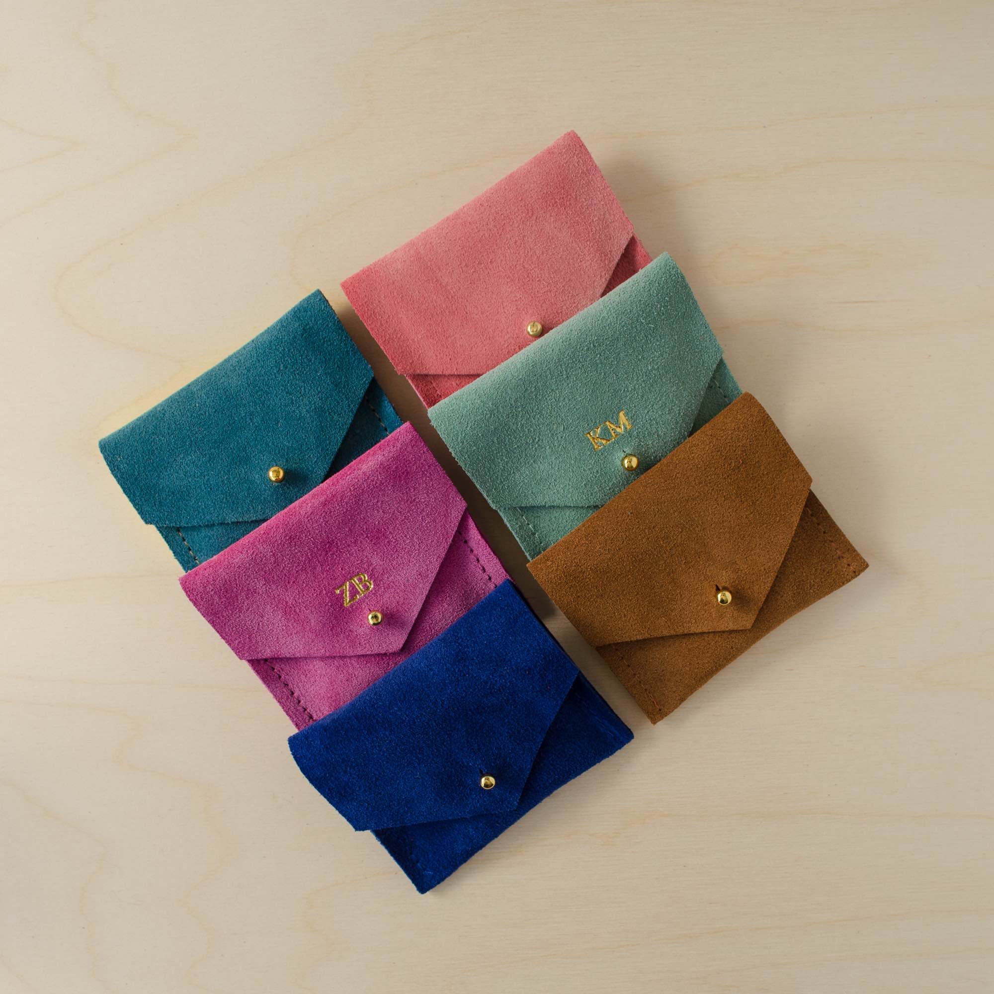 Collection of suede jewellery pouches. Teal, pink, coral, fuchsia, blue and brown options sit on a plywood table.