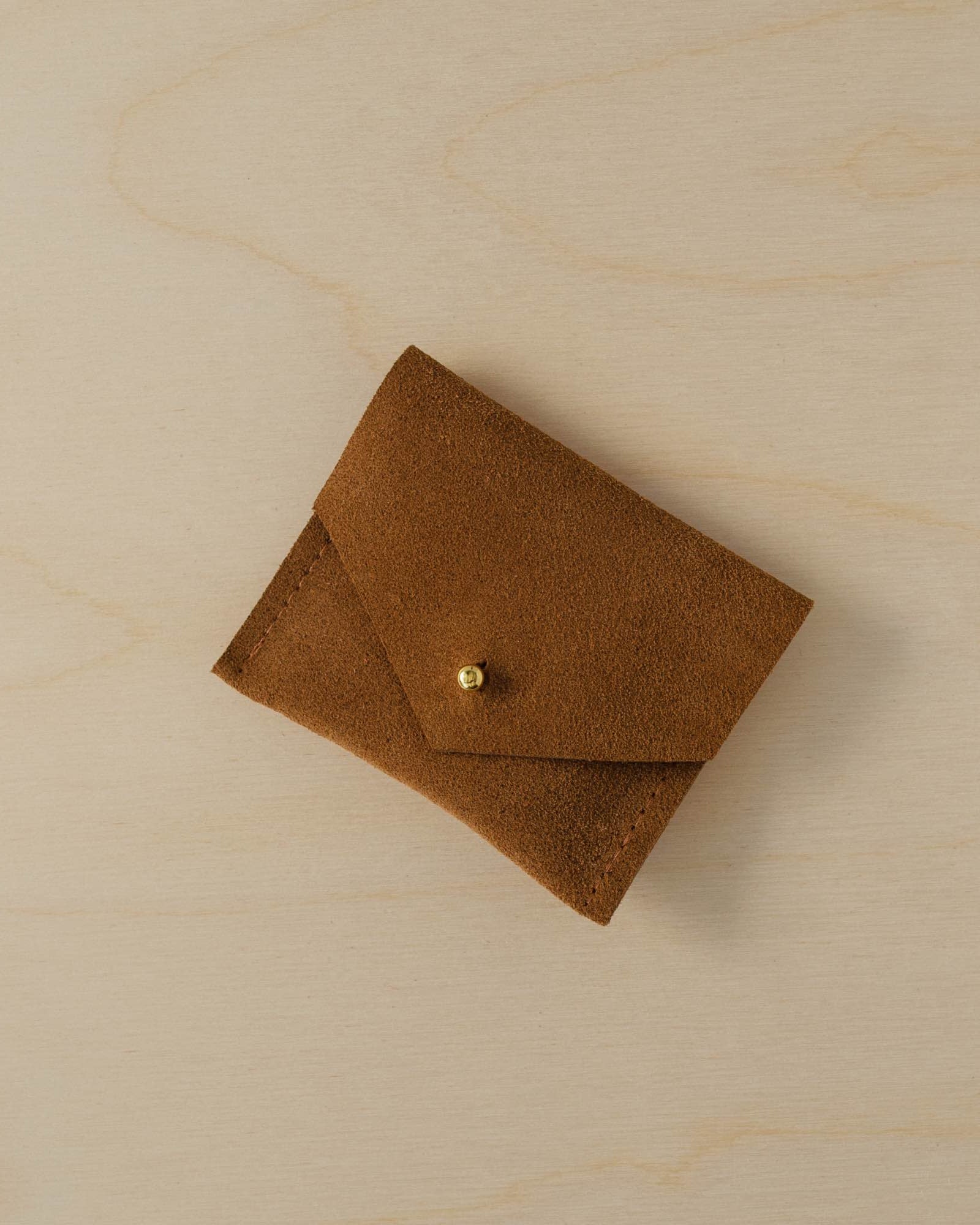 Tan brown envelope style suede jewellery pouch sits on a plywood table.