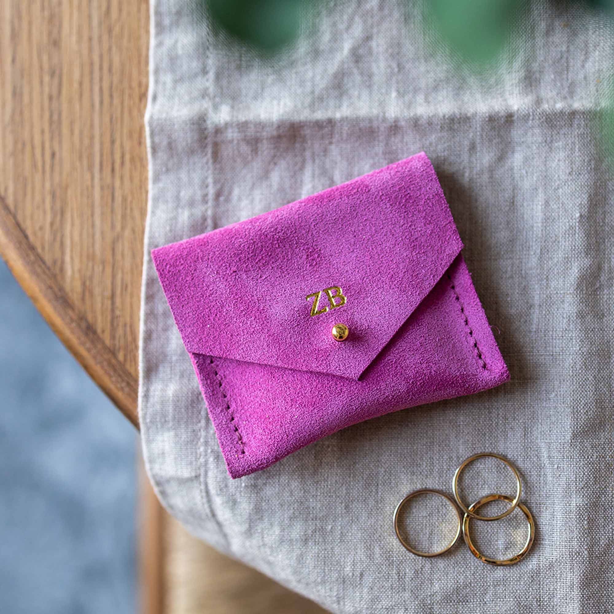 Bright fuchsia pink suede jewellery pouch sits on a desk with some silver and gold rings.