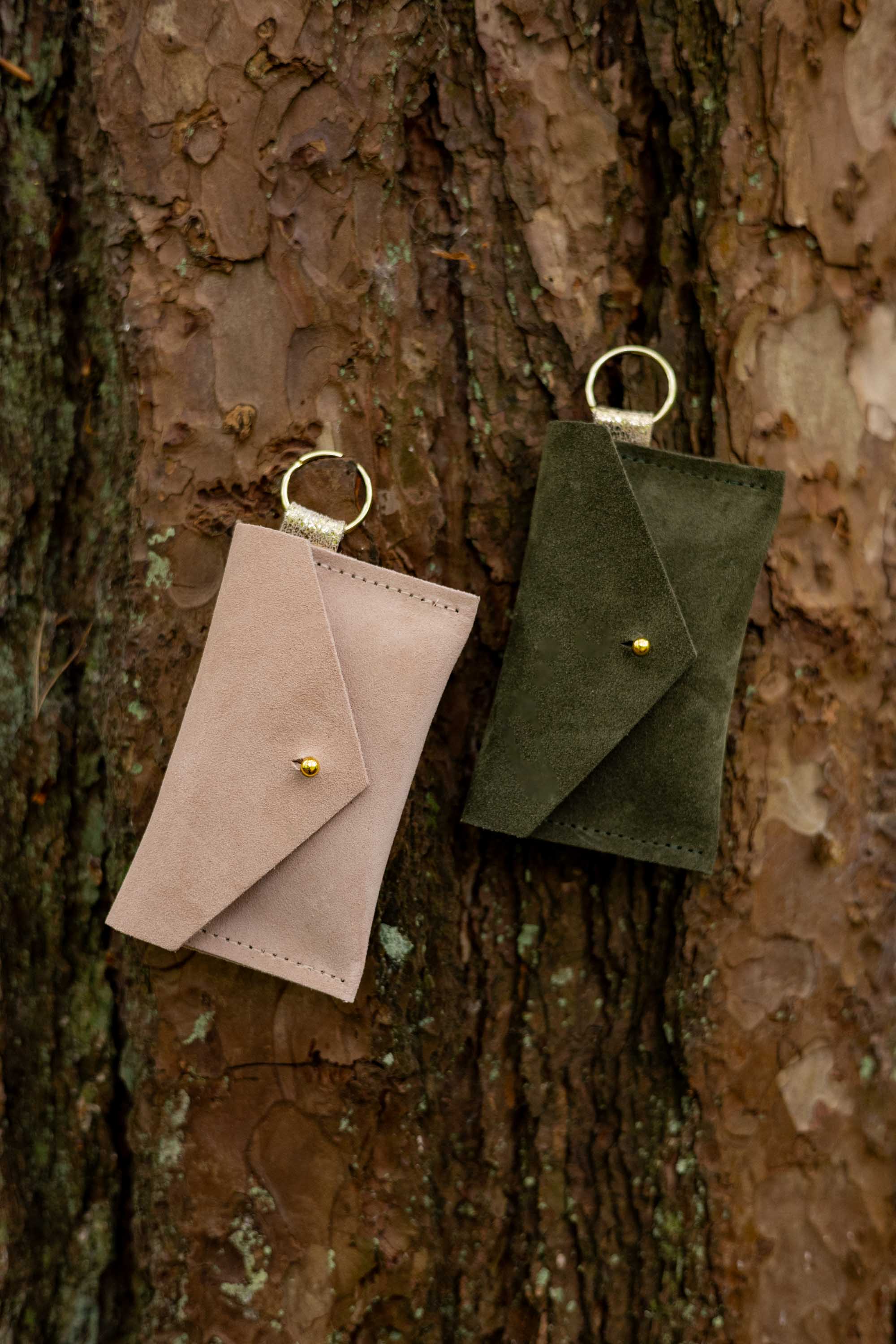 Two small suede envelope-style keyring pouches with a gold button, resting against a textured tree.