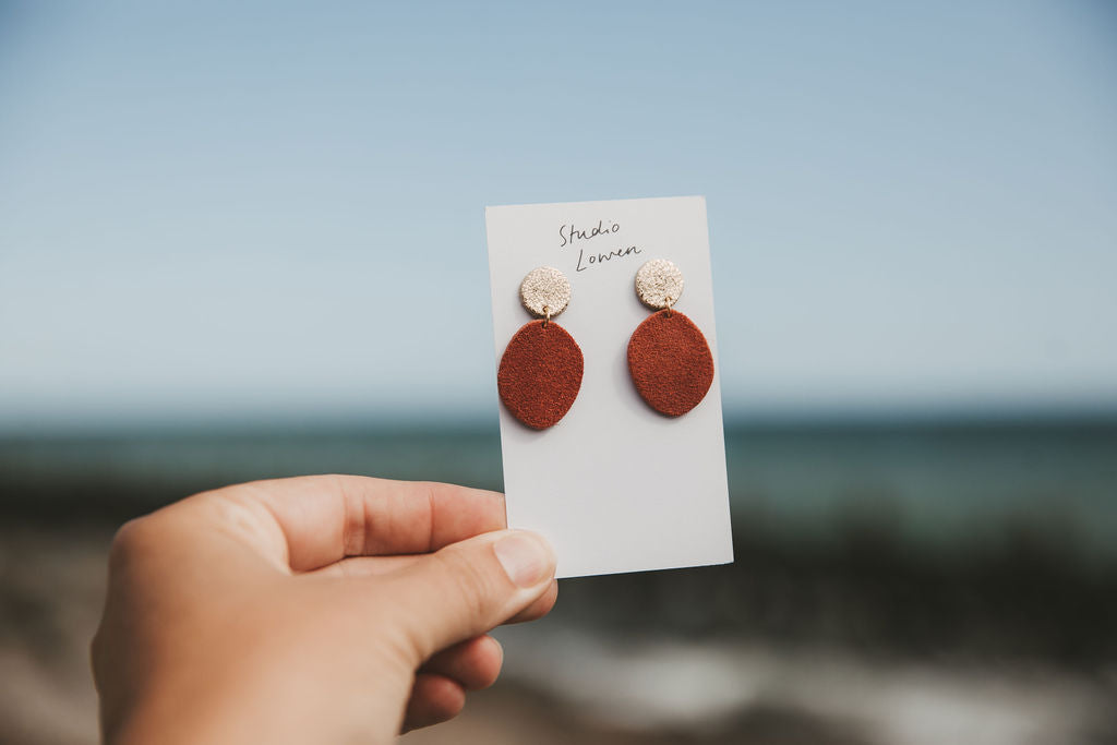 A hand holds a small white card displaying a pair of earrings against a blurred beach and sky background. The earrings have round terracotta-colored drop shapes with textured metallic gold studs above them.