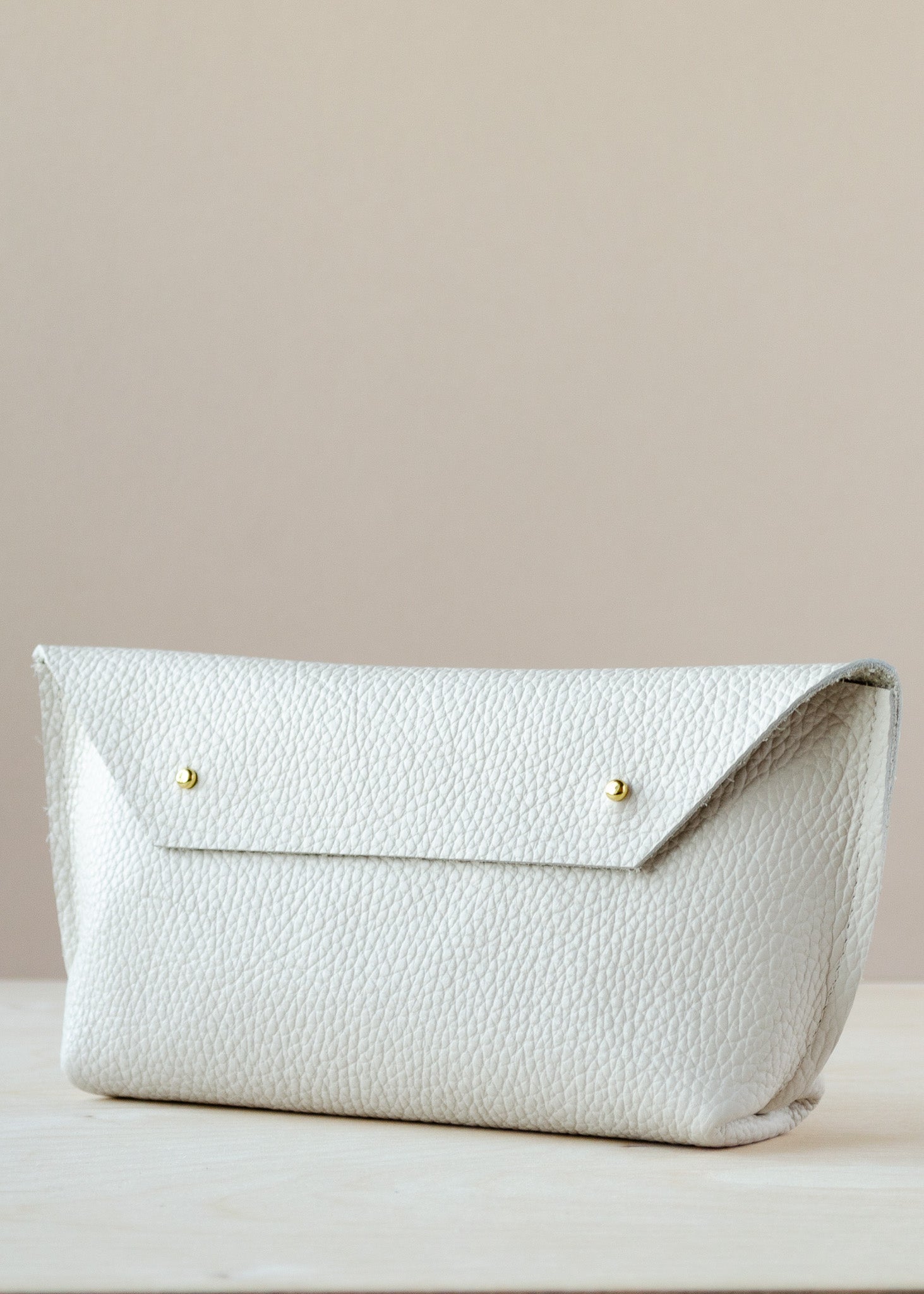 A white colored pebbled leather clutch bag with a flap closure and two small gold-tone buttons, resting on a light wooden surface against a solid beige background.