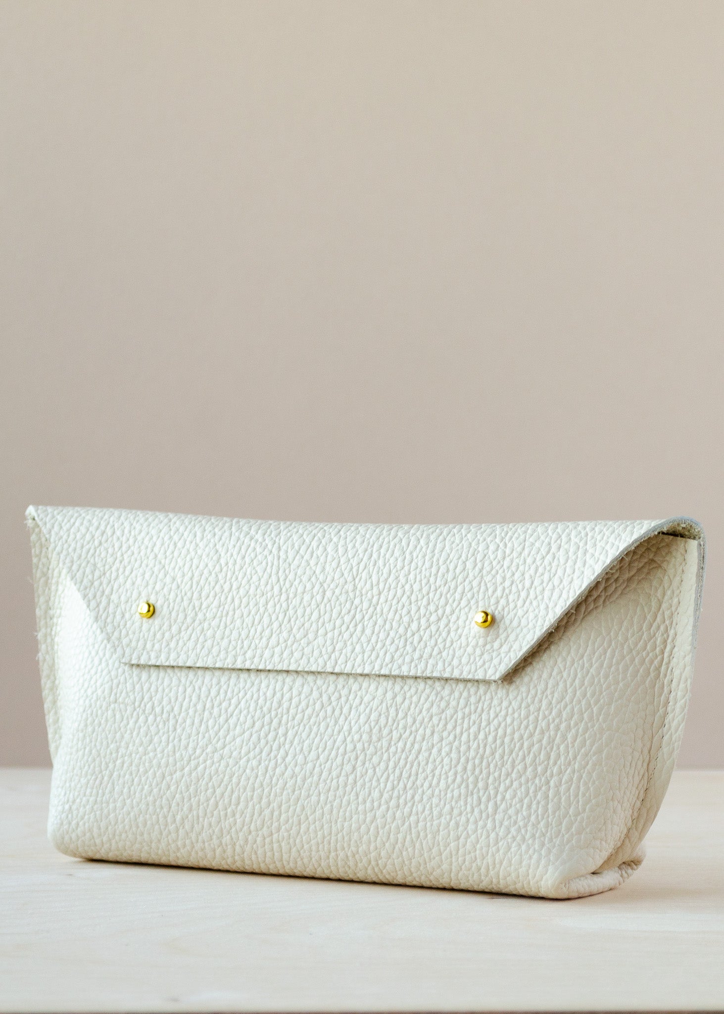 A cream-colored pebbled leather clutch bag with a flap closure and two small gold-tone metal buttons, resting on a light wooden surface against a solid beige background.