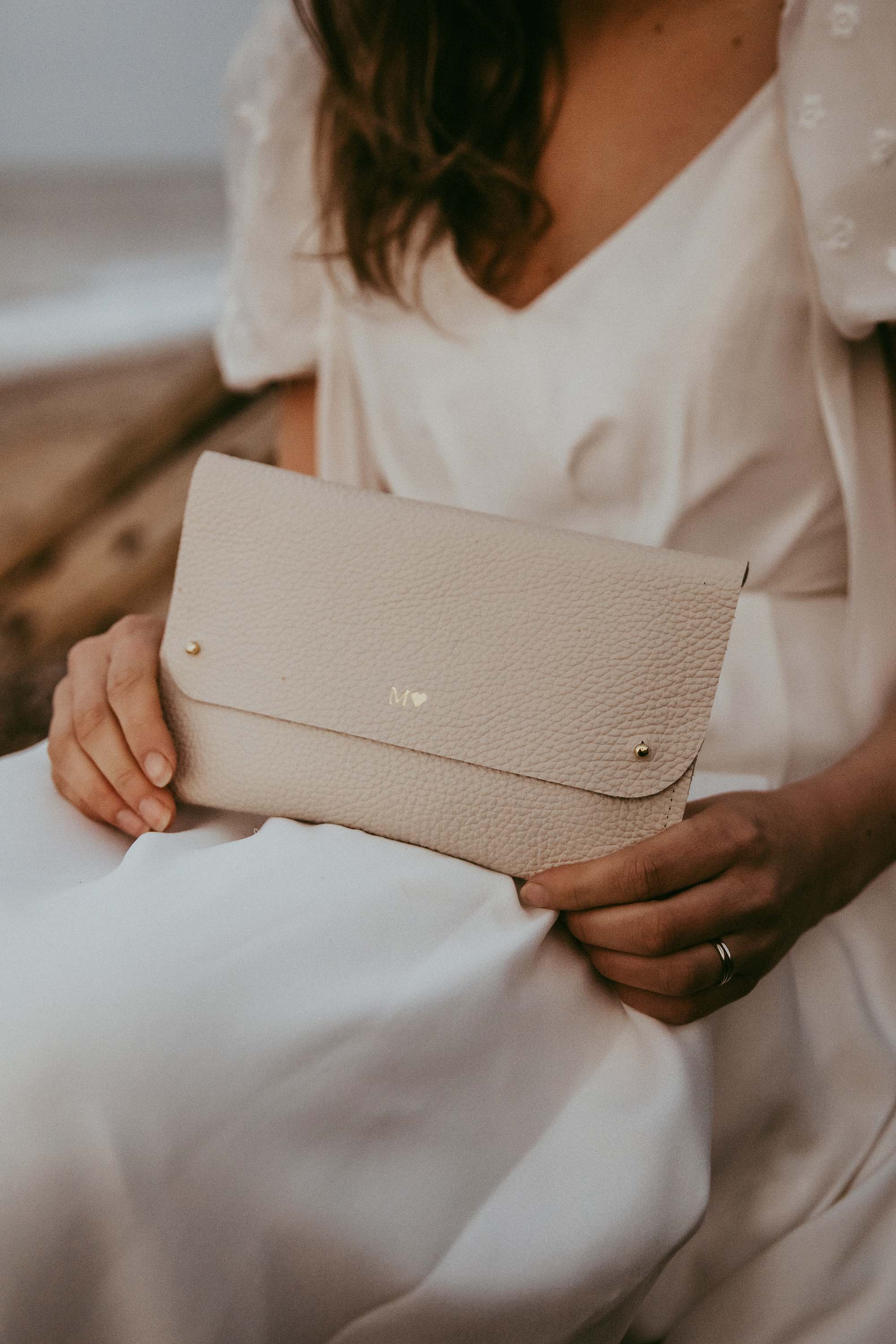 A bride in a white wedding dress holds a light beige or cream pebbled leather clutch bag with gold-tone hardware.