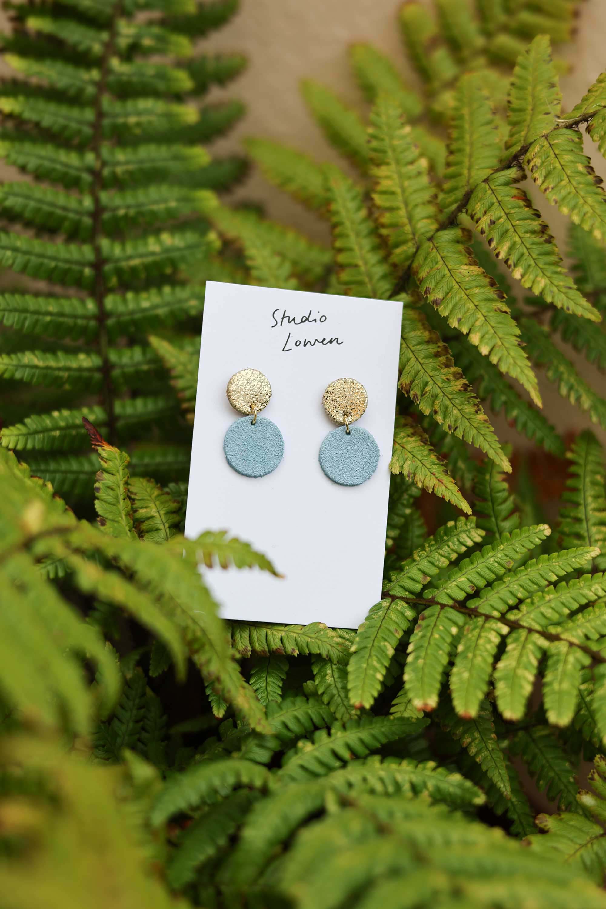 Pair of round suede and leather earrings on a card with 'Studio Lowen' branding, set against a background of green fern leaves.