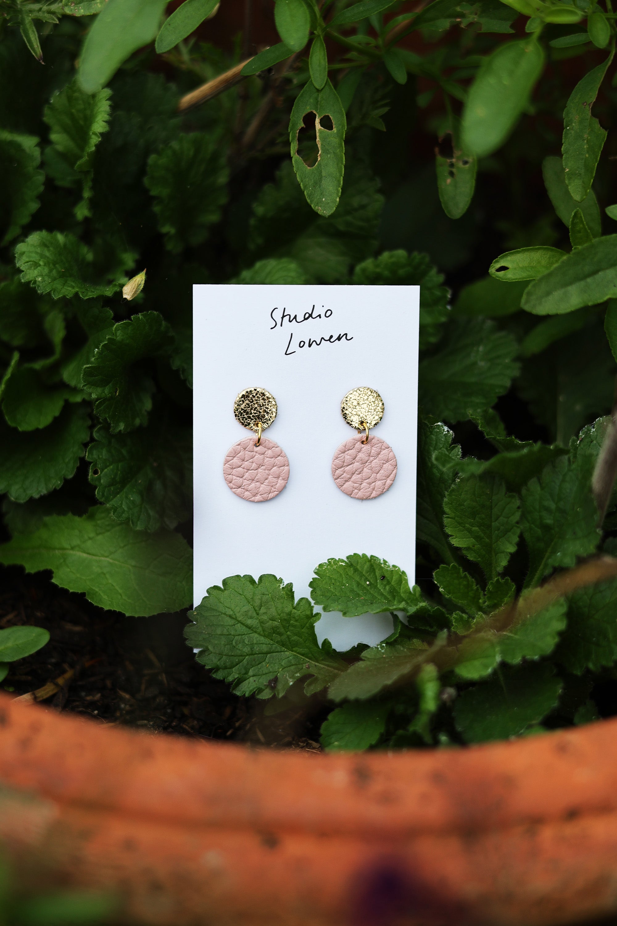 Pair of pink and gold earrings on a white card with 'Studio Lowen' branding, placed on green leaves.
