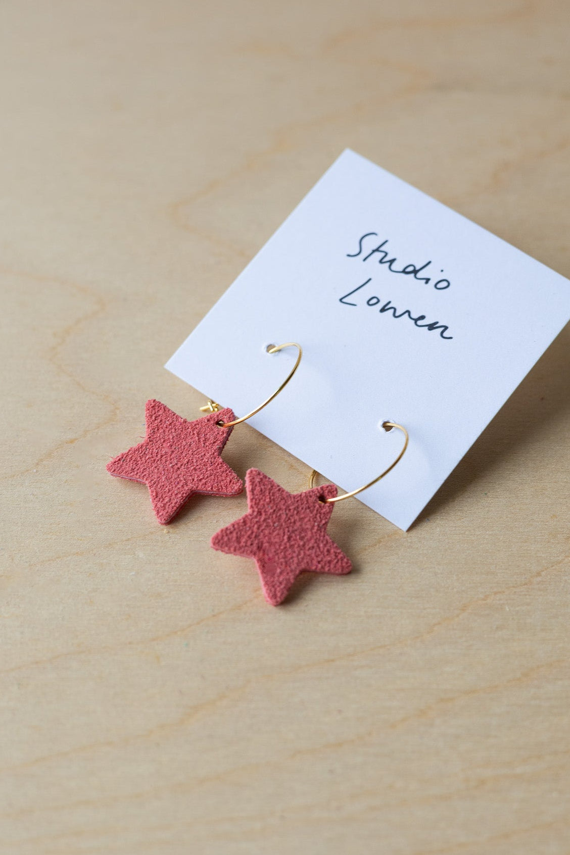 Pink star-shaped earrings on a white card with 'Studio Lowen' text, placed on a wooden surface.