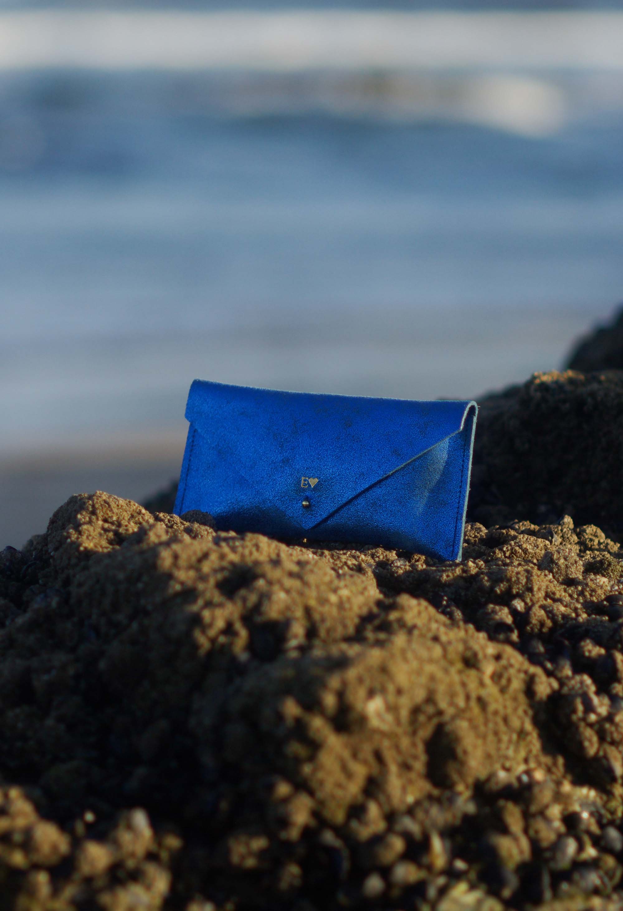 Blue leather clutch bag on a sandy beach with ocean in the background