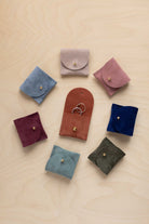 Seven small suede ring pouches in assorted colours arranged in a circle on a light wood background. The center pouch is open, holding two rings, and the others are closed with gold-tone snap buttons.