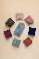 Seven small suede leather jewellery pouches in different colours arranged on a light wood background, with one blue pouch holding a gold necklace.