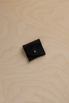 A small black suede ring pouch with a gold snap button, placed on a light wood grain surface.