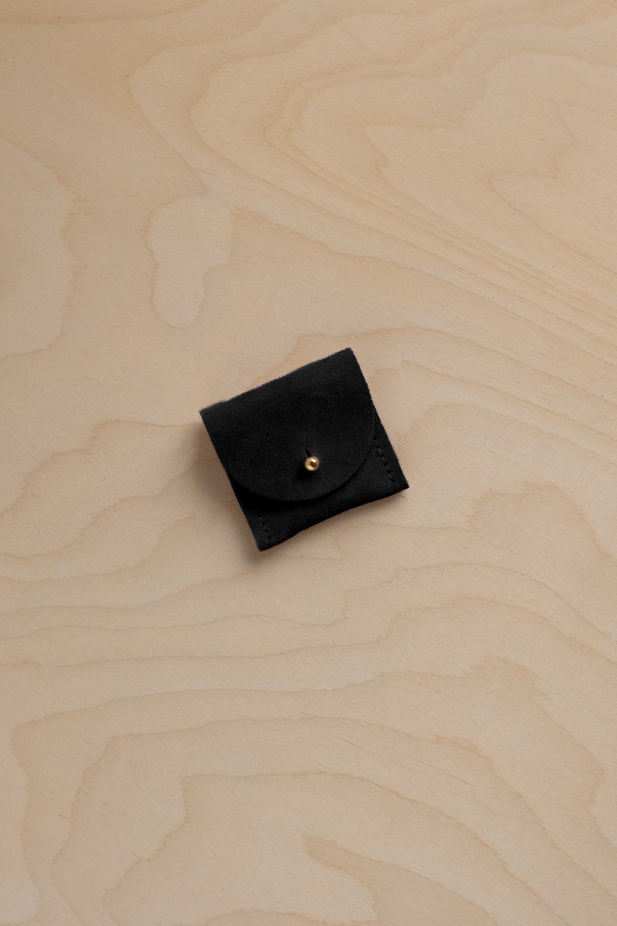 A small black suede ring pouch with a gold snap button, placed on a light wood grain surface.
