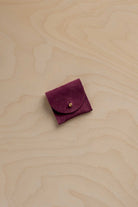 A small burgundy suede pouch with a gold button, placed on a light wood grain background.