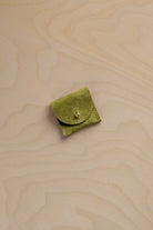 A small light olive green suede leather pouch with a button closure, placed on a light beige wood-grain surface.
