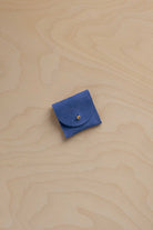 A small cobalt blue leather pouch with a flap closure and a tiny gold button, placed on a light wood grain surface.