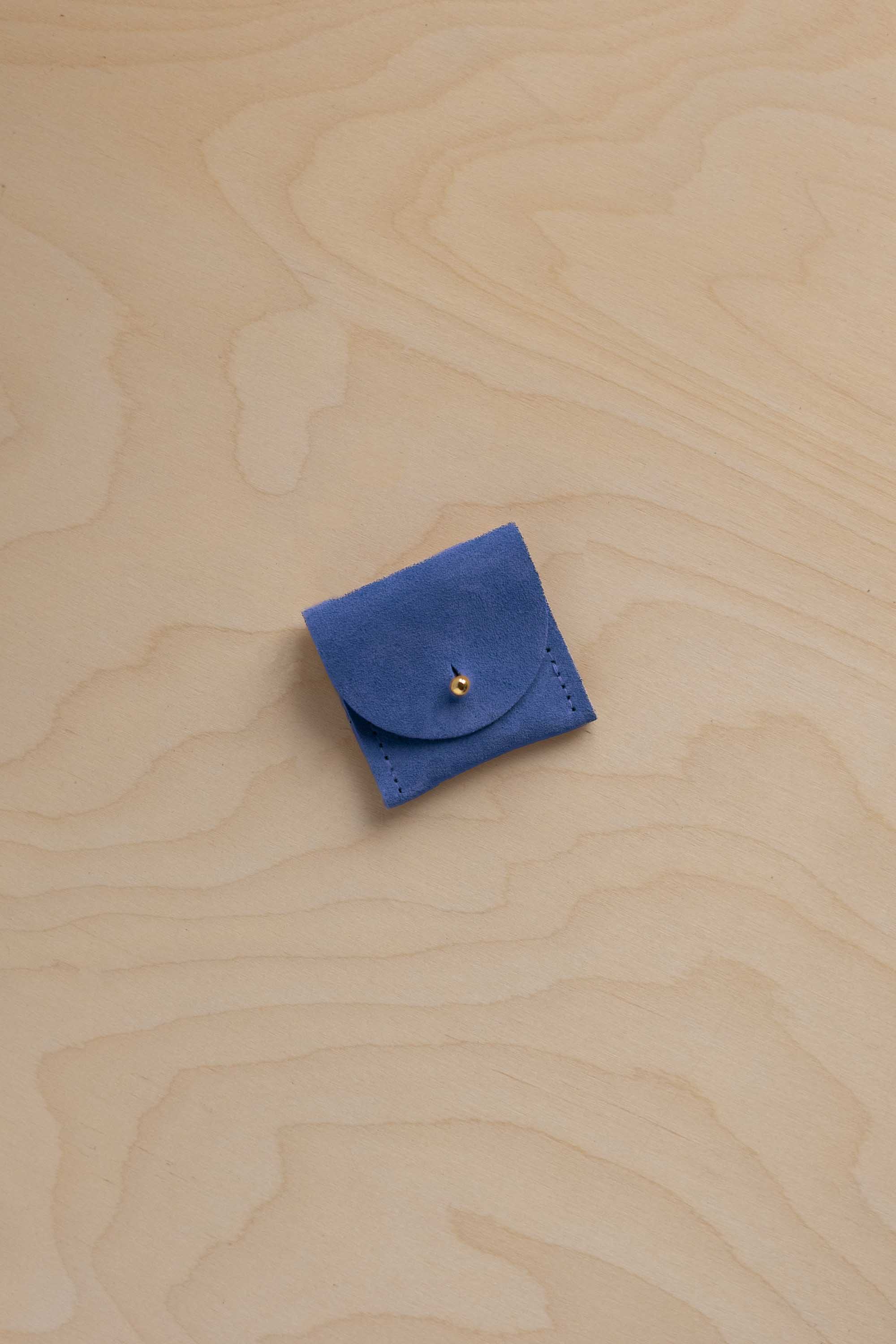 A small cobalt blue leather pouch with a flap closure and a tiny gold button, placed on a light wood grain surface.