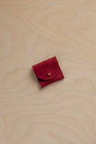 A small deep red suede leather pouch with a gold button closure, placed on a light beige wood grain background.