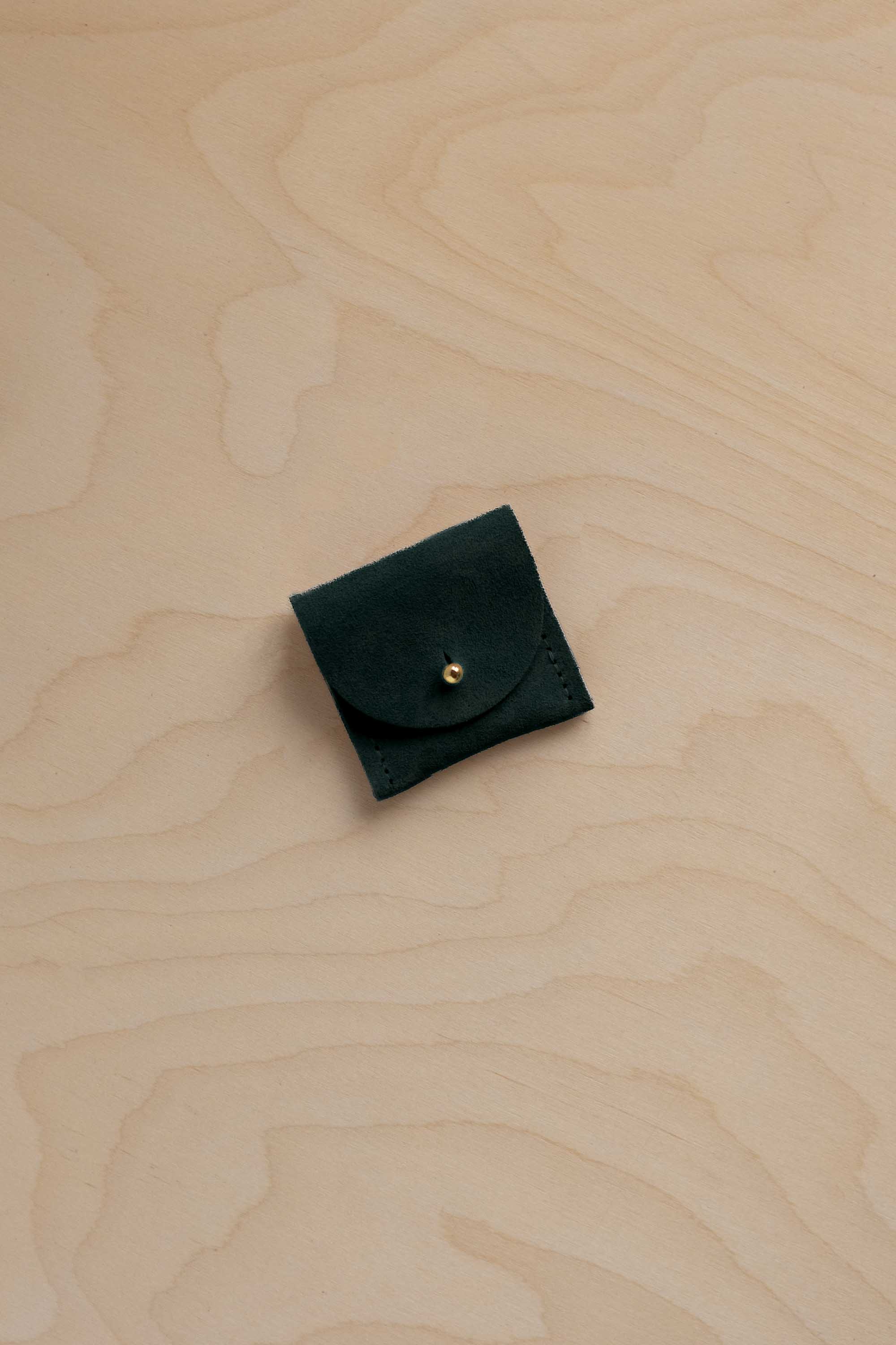 A small dark forest green suede leather pouch with a flap closure and a gold button, placed on a light wood grain surface.