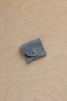 A small blue gray suede ring pouch with a flap closure and a tiny gold button, placed on a light wood grain surface.