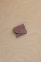 A small dusty mauve purple suede leather pouch with a flap closure and a tiny gold fastening, placed on a light wood grain surface.