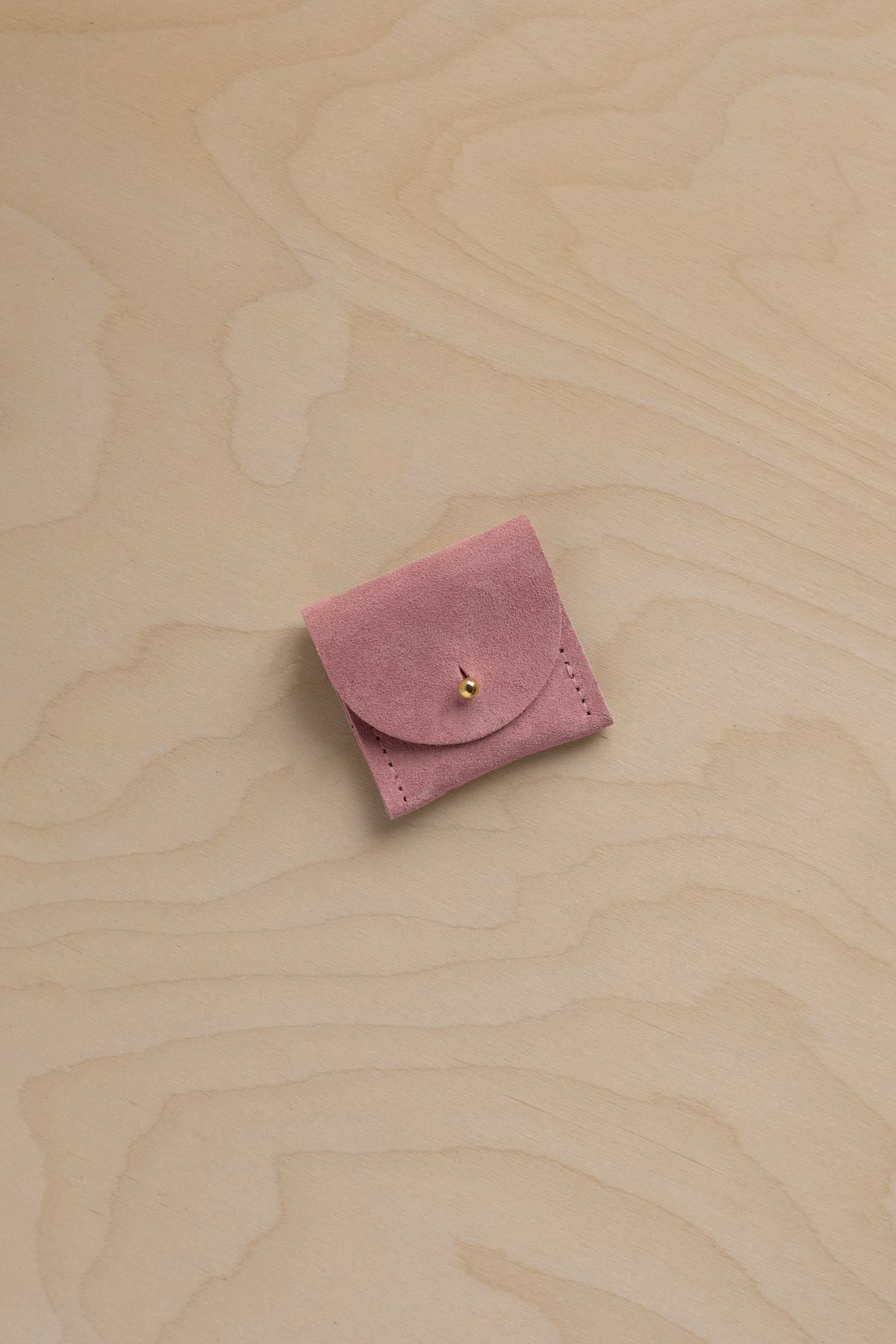 A small dusty pink suede jewelry pouch with a flap closure and a tiny gold bead accent, resting on a light wood grain surface.