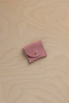 A small dusty pink suede jewelry pouch with a flap closure and a tiny gold bead accent, resting on a light wood grain surface.