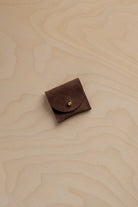 A small dark brown suede leather pouch with a flap closure and a gold snap button, placed on a light wood grain surface.
