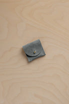 A small sage green suede pouch with a flap closure and a gold fastening, placed on a light wood grain surface.