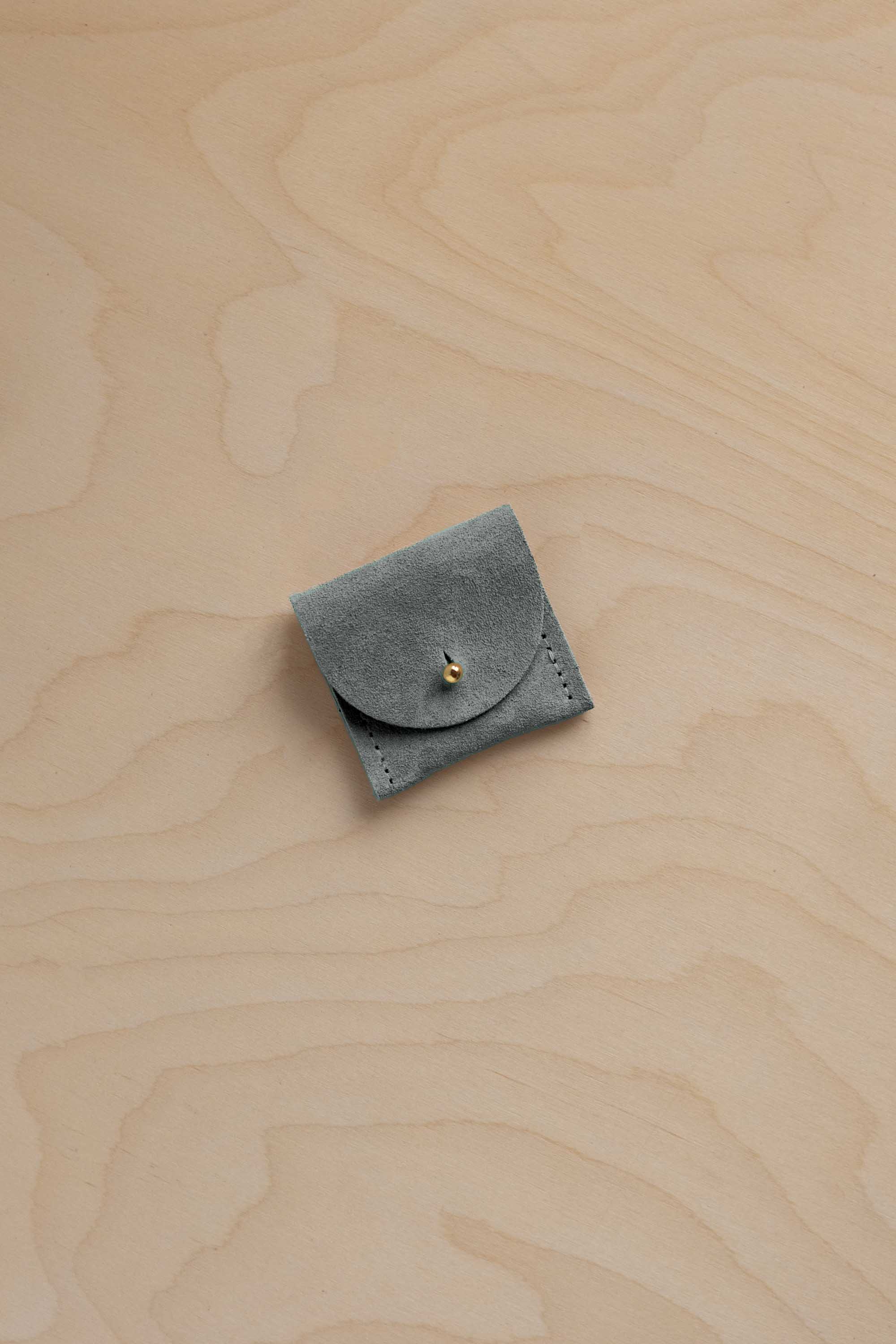 A small sage green suede pouch with a flap closure and a gold fastening, placed on a light wood grain surface.