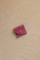 A small rose pink suede pouch with a gold button closure, placed on a light beige wood grain background.